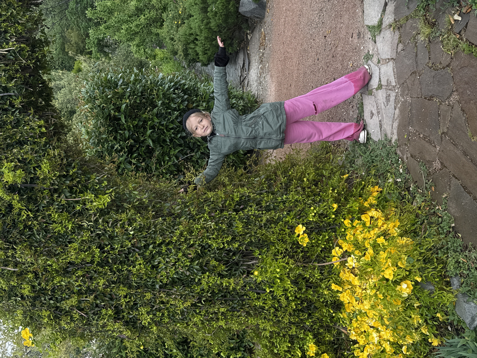 A young girl stands outside amidst lush vegetation. She is wearing a green jacket, pink pants, a black hat, and gloves. Her arms are outstretched. She stands on a path constructed of stone and gravel. In the background is a dense growth of green bushes, some with yellow flowers. The location is identified as Unknown, Unknown.