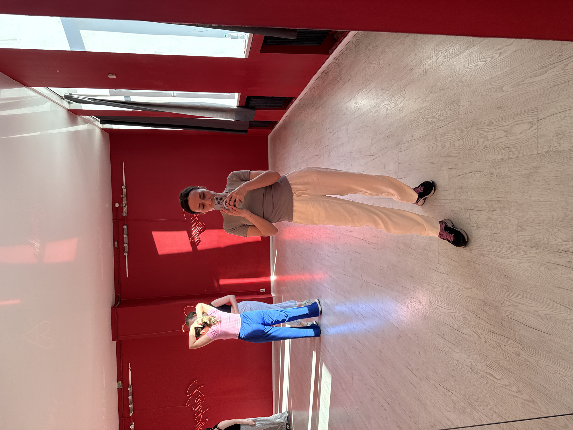 A woman in a gray t-shirt and light-colored pants takes a selfie in what appears to be a dance studio with red walls and a white floor. She is holding her phone up and looking into the camera. Reflected in the mirror are other people in dance attire, as well as a red neon sign. The location is Saint Petersburg, Russia, based on the Cyrillic lettering of the neon sign reflected in the mirror.