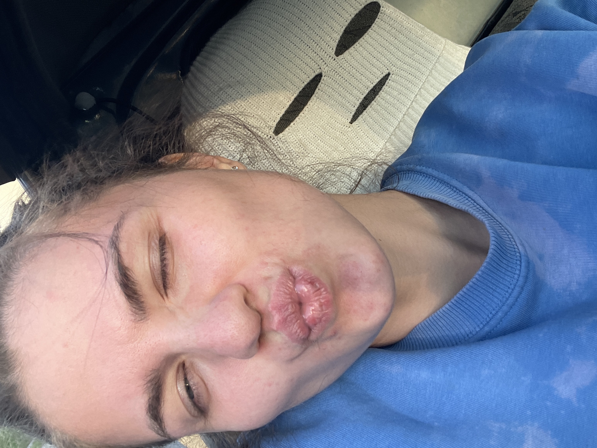 A selfie of a fair-skinned person with brown hair and eyes, wearing a blue sweatshirt. The person is making a kissing face. A knitted cushion with dark vertical stripes is partially visible in the background, along with a dark object. The location is not identifiable as Molokovo, Russia.
