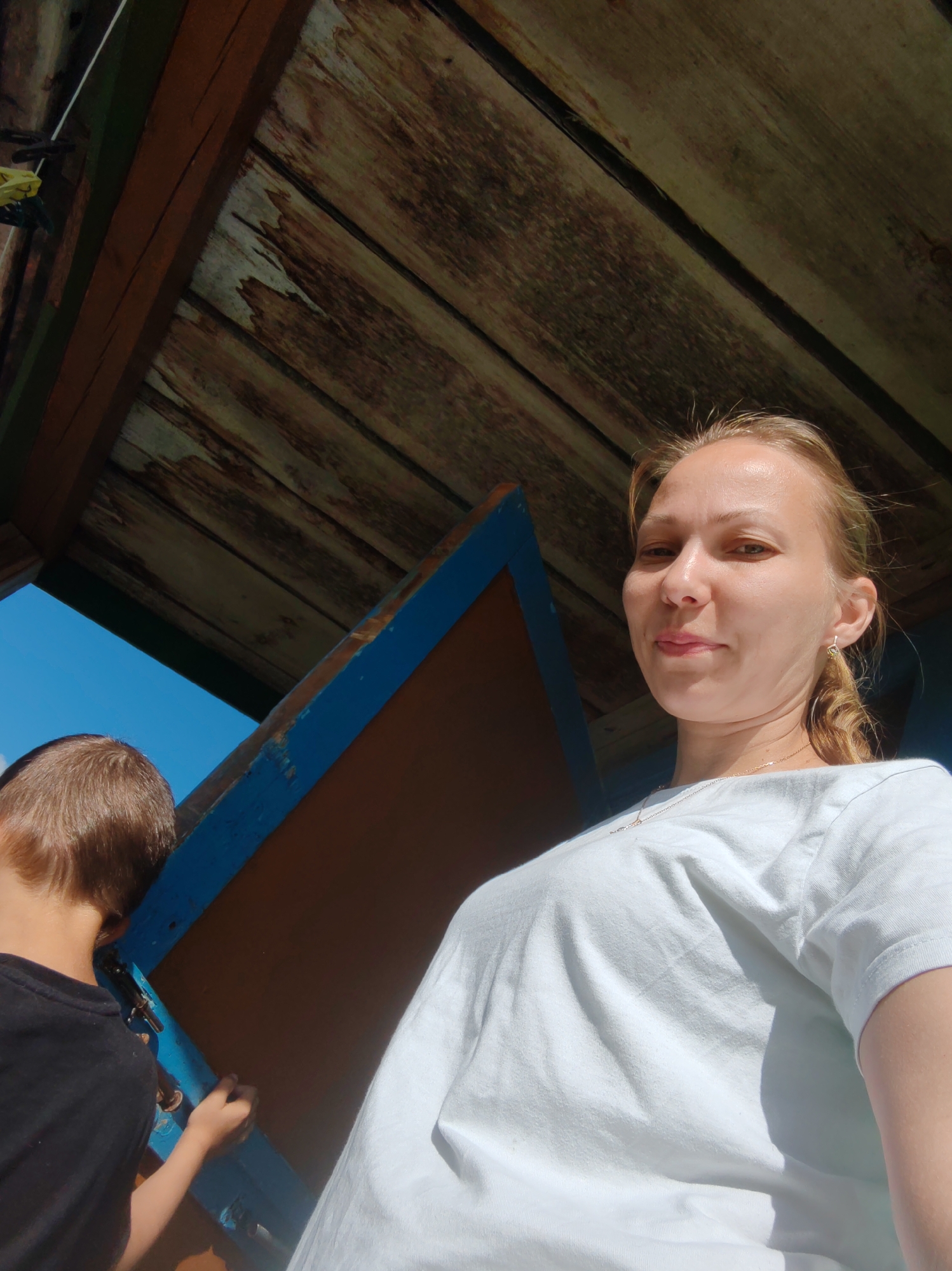 A low-angle, eye-level, waist-up shot captures a woman with blonde hair pulled back, wearing a white t-shirt and a thin necklace. She is looking directly at the camera with a slight smile. To her left, a young boy with short brown hair is seen from behind, wearing a black t-shirt and engaged in an action with a blue and brown structure. The background consists of weathered wooden planks forming a ceiling or overhead structure, with a clear blue sky visible through an opening. The scene appears to be outdoors, possibly in a park or a similar public space.