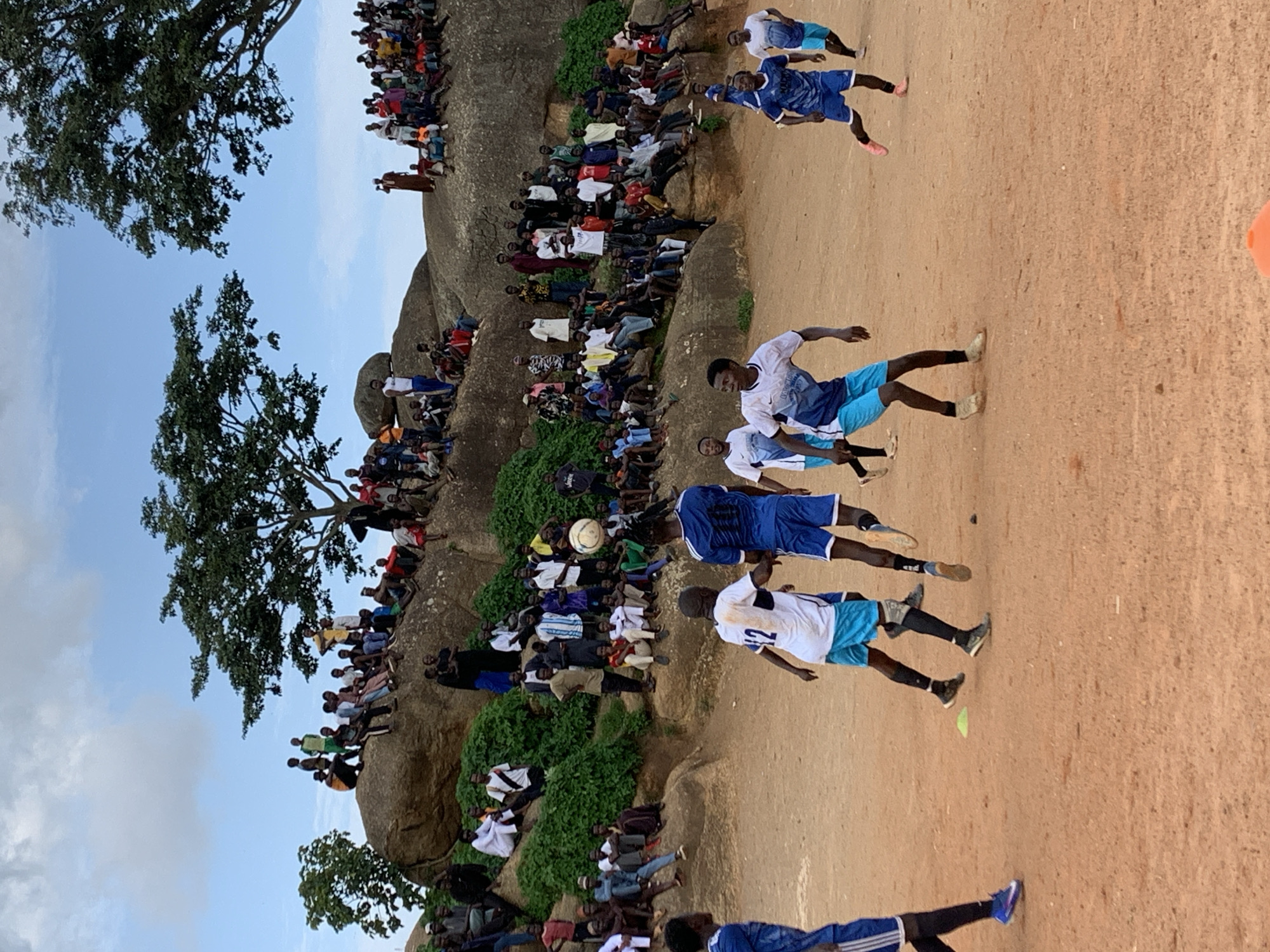 The image depicts an outdoor football match taking place on a dirt field. The playing area is sandy and uneven. In the foreground, four young men in football attire are actively engaged in the game, with one player in the center kicking a ball. The players are wearing blue and white jerseys with blue shorts and black socks.
The background is dominated by a rocky, elevated terrain where a large crowd of spectators is gathered. These spectators are seated and standing on the rocks, observing the game. Their clothing varies, with many individuals wearing casual attire. Two large trees with dense foliage are visible on the rocks, providing some shade. The sky above is partly cloudy with a mix of blue sky and white clouds.
The scene suggests a community event or a local sporting competition, likely held in an area with distinct rock formations, which are characteristic of certain parts of Jos, Nigeria. The overall atmosphere appears lively and engaged, with the spectators keenly following the football match.