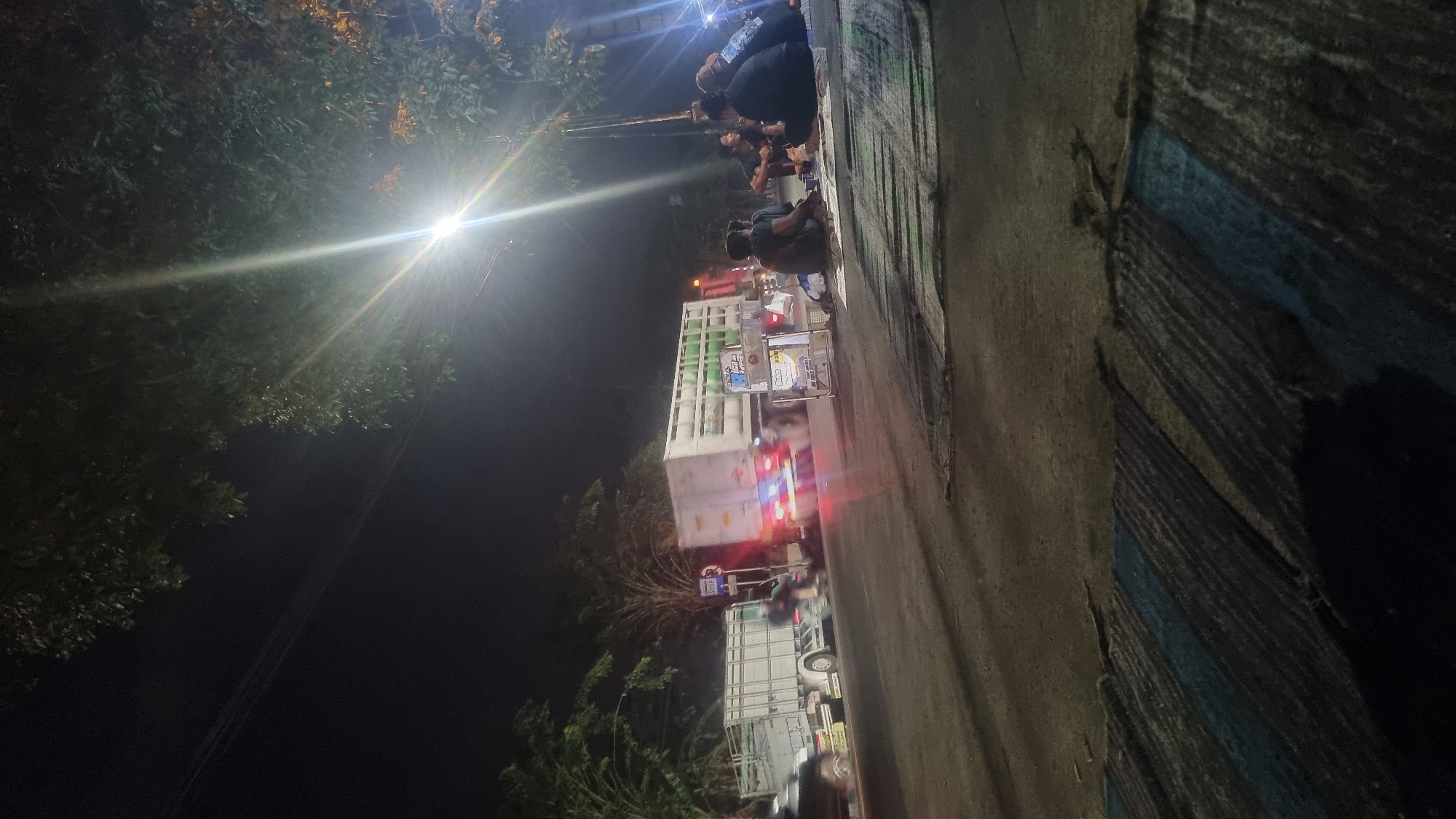 At night in Jotangan, Indonesia, a group of people are sitting on the ground next to a road. A large truck is driving by on the road, and another truck is parked on the side of the road. There are trees in the background, and a street light is shining down on the scene. The people appear to be socializing, and some of them are looking at their phones. The scene is dimly lit, and the overall mood is relaxed.