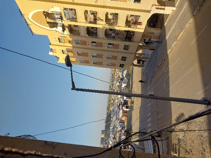 An elevated view captures a street scene in Khan Yunis, Palestinian Territory, under a clear blue sky. In the immediate foreground on the left, a portion of a light-colored building structure with attached wires is visible. A paved, two-lane road with a faded white crosswalk and a white turn arrow occupies the midground. A tall, grey streetlamp with overhead wires extends into the frame from the center-left.
To the right, a multi-story, light yellow residential building with brown window accents and decorative arched architectural elements on its upper facade stands prominently. Several balconies on the building's right side display various items of clothing, including red, white, and blue fabrics, hanging to dry. The ground floor features a series of arches forming an open-air space, with a dark-colored vehicle parked beneath one arch on the far right. Approximately 5-7 individuals are present on the sidewalk and under the building's arches, with one person walking on the road further back.
Beyond the street and a dusty, unpaved area, the background reveals a large, dense encampment composed of numerous white and grey makeshift shelters, tents, and rudimentary structures. Figures are discernible moving within this encampment. In the far distance, a large, light-colored cylindrical structure and a fortification-like building are visible on an elevated landform, backed by distant hills. The scene is illuminated by direct sunlight, casting distinct shadows across the street.
