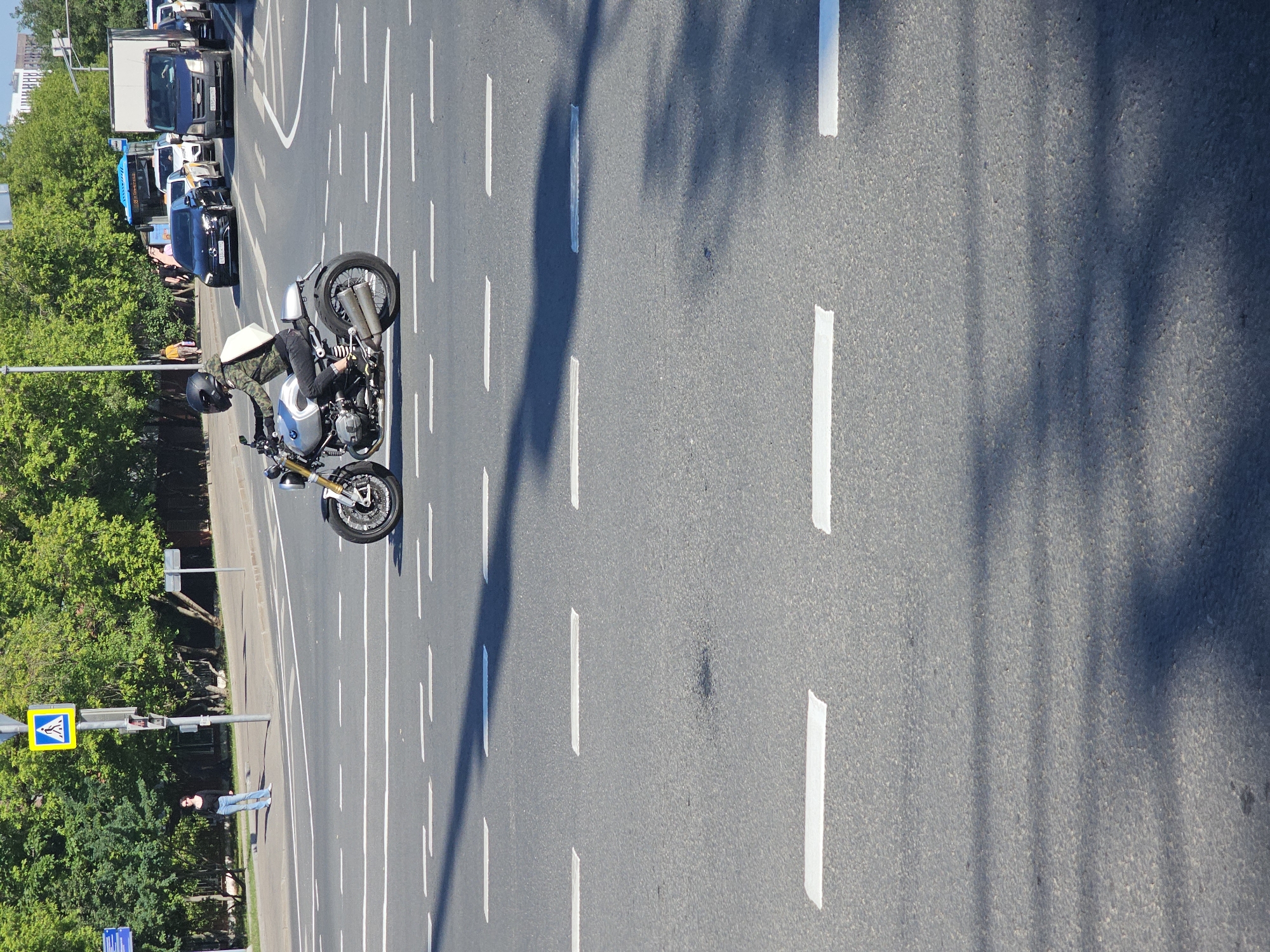 This high-angle, downward-looking photograph captures a sunny urban or suburban road scene in Moscow, Russia. The image is oriented with the multi-lane asphalt road running lengthwise from bottom to top.
In the foreground/midground, a motorcyclist is riding or stationary on a distinct silver and black motorcycle, which appears to be a BMW R nineT or a similar scrambler-style model, identifiable by its spoked wheels and visible exhaust system. The rider is equipped with a black full-face helmet, a camouflage-patterned jacket, and dark trousers.
The road features numerous dashed white lane markings, indicating multiple lanes of traffic. To the left of the road, there is a sidewalk or shoulder lined with abundant green trees. A yellow pedestrian crossing sign, featuring a blue border and black walking figures, is visible on a pole near the trees. Further along the road in the background, a queue of vehicles, including several cars and at least two trucks, suggests active traffic.
Distinct, elongated shadows are cast across the asphalt from the right side of the frame, indicating bright sunlight and a clear sky, consistent with mid-day or afternoon conditions. No specific textual information is clearly readable on vehicle license plates.