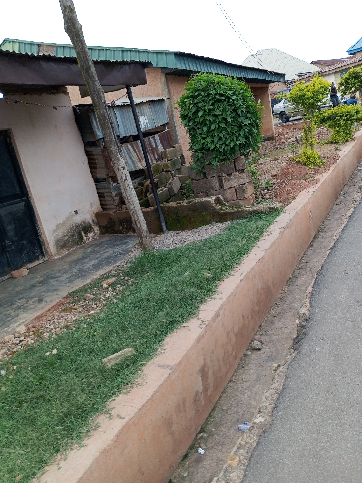 The image depicts an outdoor scene in Gura Top, Nigeria, showing a street-level view of a building and its surroundings. The building has a corrugated metal roof, with sections of rust-colored metal and corrugated aluminum siding visible. A large, dark door is present on the left side of the building, with a light-colored, weathered wall. Adjacent to the building, a wooden pole stands vertically, leaning slightly. Piles of concrete blocks are stacked in front of the building. A thick, green bush is situated to the right of the building's entrance. In the background, several small trees with light green foliage are planted along a concrete barrier. A light-colored sedan is parked on the street, with two individuals standing near it, one of whom is looking away from the camera. The street itself appears to be paved, with a concrete drainage channel running alongside it. The ground between the building and the drainage channel is covered in short green grass, interspersed with scattered pebbles and some debris. The overall impression is of a slightly unkempt urban environment.