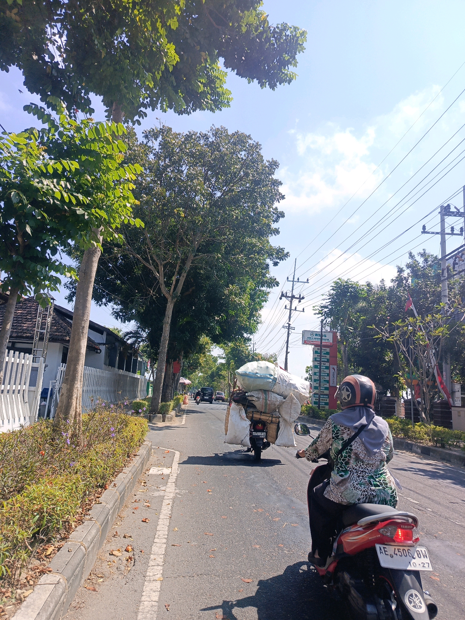 The image depicts a street scene in the city of Tambran, Indonesia. In the foreground, a person is riding a red Yamaha scooter with the license plate AE 4506 BW. They are wearing a helmet and a patterned shirt. In front of this scooter, another motorbike is visible, heavily laden with large white sacks. The road is paved, with a curb and a sidewalk lined with green bushes and trees. Houses with tiled roofs and white fences are visible on the left side of the street. Utility poles with numerous wires are present on the right side of the road, along with a red sign that appears to be for a hospital. The sky is blue with some scattered clouds, and the sunlight indicates it is daytime. The overall impression is that of a busy street with ongoing activity.
