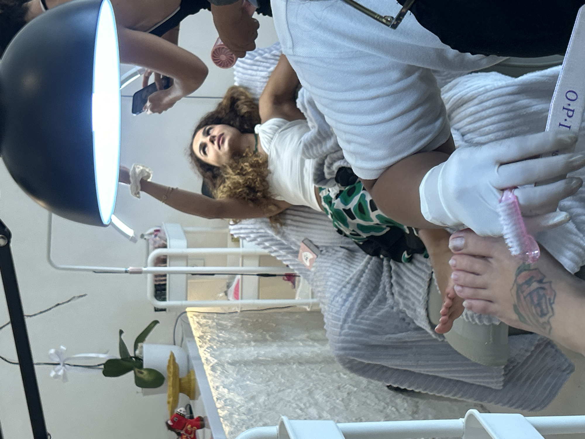 In North Kuta, Indonesia, a person is receiving a pedicure. They are reclined in a chair, with their feet propped up. One foot is closer to the camera and shows a rose tattoo on the ankle and is being worked on by someone wearing white gloves and holding a nail file. The other foot is further away and is also receiving attention. A bright lamp is positioned above the seated person, who is looking up. Other people and salon equipment are visible in the background, including a shelf unit with various items and a plant in a pot.