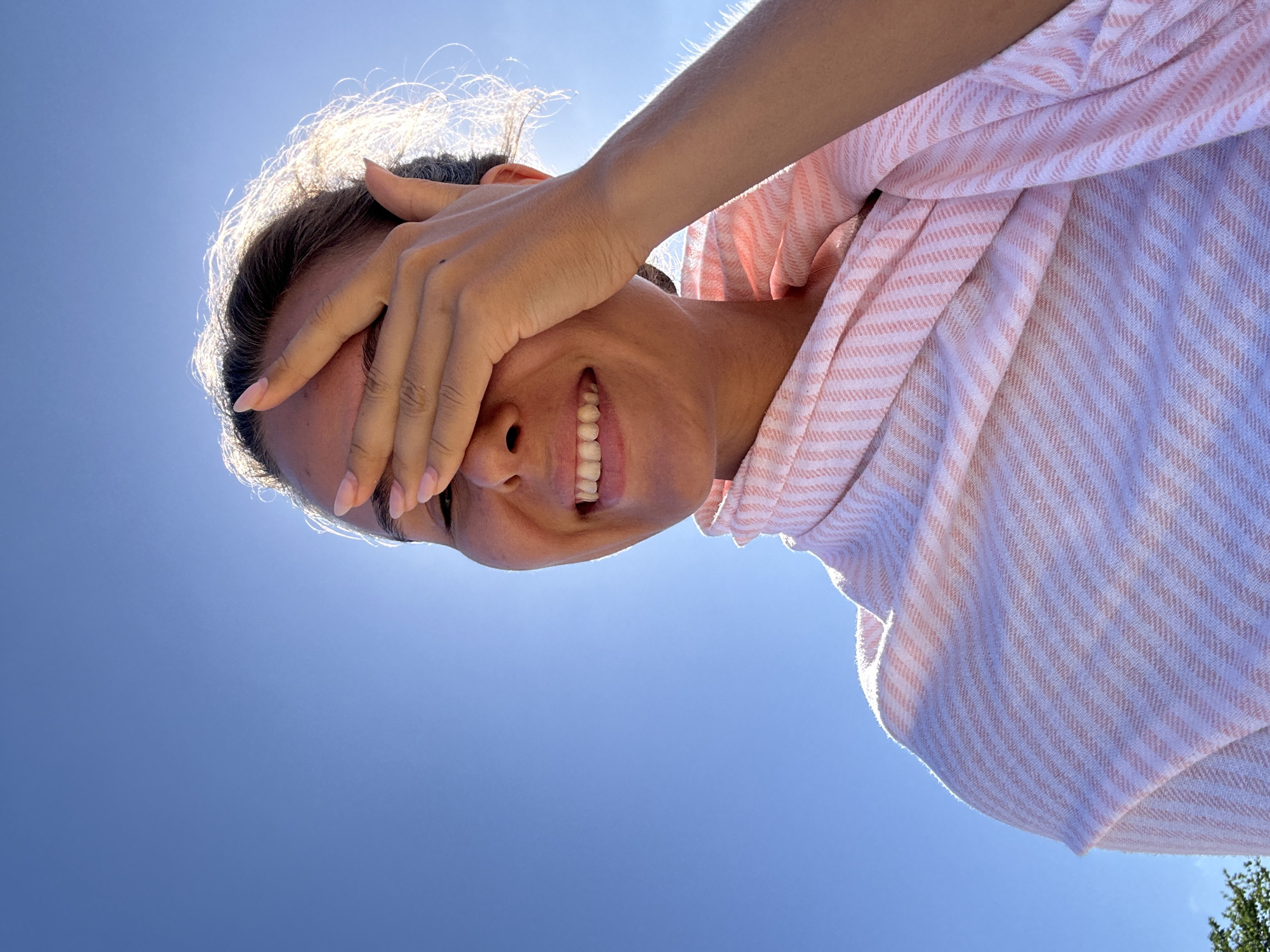 In the city of Kuta Selatan, Indonesia, a young woman with brown hair is captured in a close-up, low-angle shot, looking up towards a clear blue sky. Her right hand is raised, covering her eyes as she smiles, revealing her teeth. She is wrapped in a pink and white striped fabric. The sun is shining brightly, creating a lens flare effect and highlighting the edges of her hair. The background consists solely of the bright, cloudless sky. A hint of greenery is visible at the bottom left edge of the frame, suggesting an outdoor natural setting.
