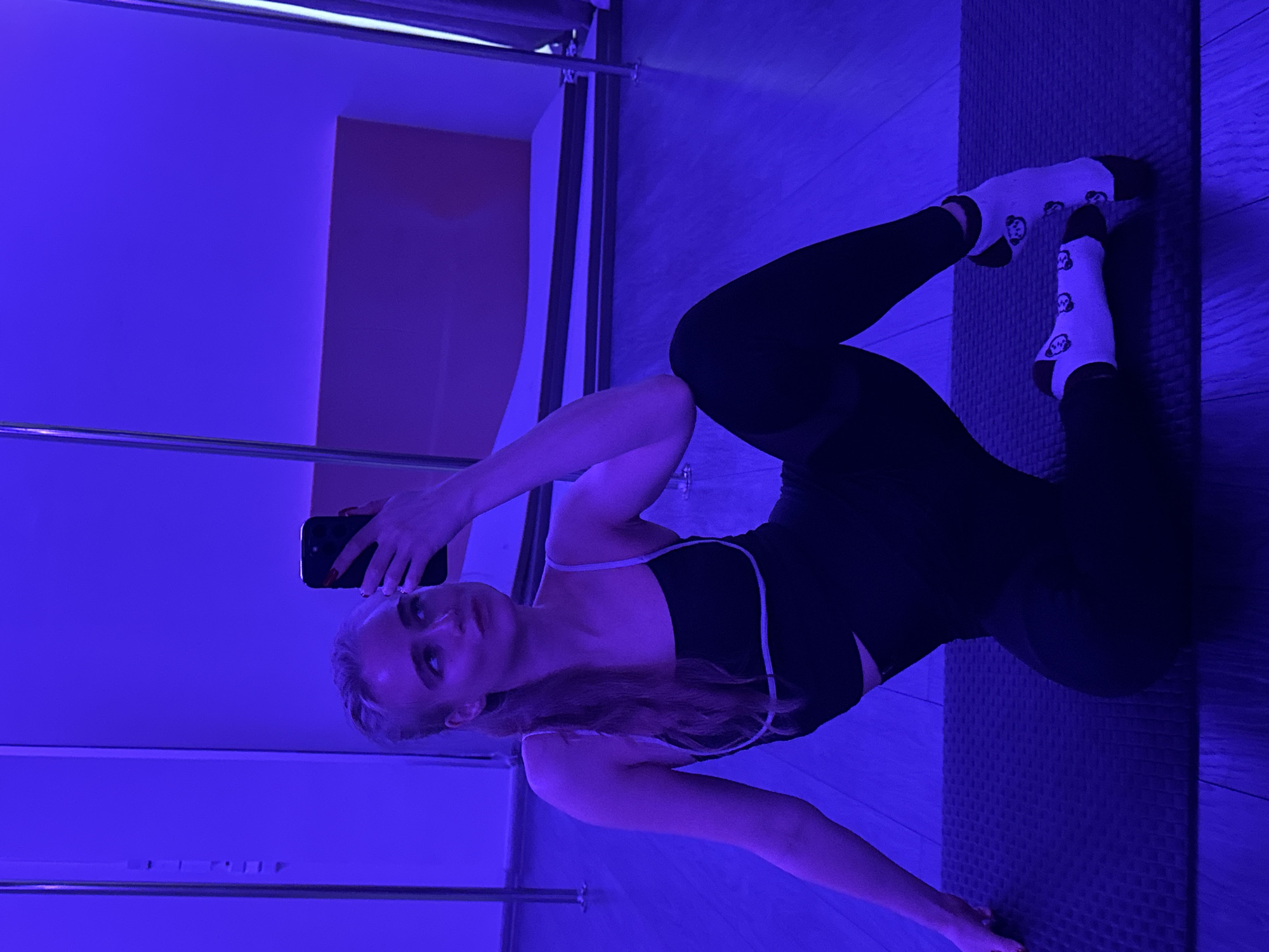 A young, fair-skinned woman with long, light-colored hair is positioned in what appears to be a mirrored room. She is dressed in a black workout outfit with white straps and black leggings, and she is wearing white socks decorated with small, repeated images. The woman is lying on a textured gray mat. Her body is angled towards the camera as she holds up a black smartphone in her right hand, seemingly taking a selfie. The room is lit with a blue hue, which obscures some details and casts strong shadows. Behind her, a partially visible shelving unit with a pink panel can be seen in the mirror’s reflection. The floor is a light-colored wood, and the room appears to be a fitness or yoga studio, though the exact location in an unknown city and state remains unspecified.