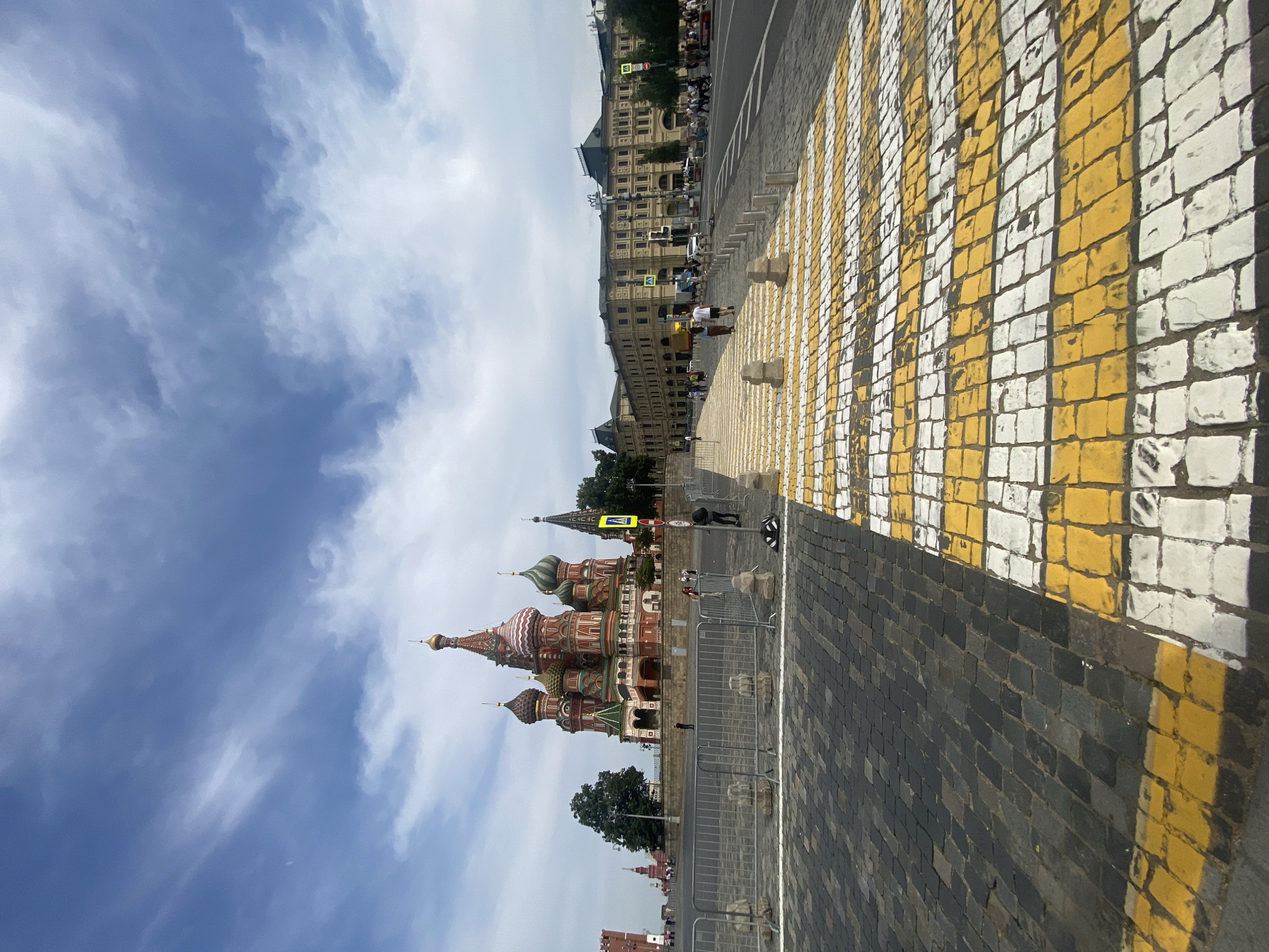 The image depicts Red Square in Moscow, Russia. In the foreground, a prominent yellow and white striped pedestrian crossing is laid out on a cobblestone surface. Beyond the crossing, a grey metal barricade runs horizontally across the scene, separating the pedestrian area from the square. In the middle ground, the iconic St. Basil's Cathedral, with its colorful onion domes, stands majestically. To the right of the cathedral, a long, yellow building with a dark roof, likely a part of the GUM department store, stretches across the background. Several people are visible in the middle and background, walking across the square and near the buildings. The sky is partly cloudy with a mix of blue sky and white clouds. The overall environment appears to be an open public space with historical architecture.