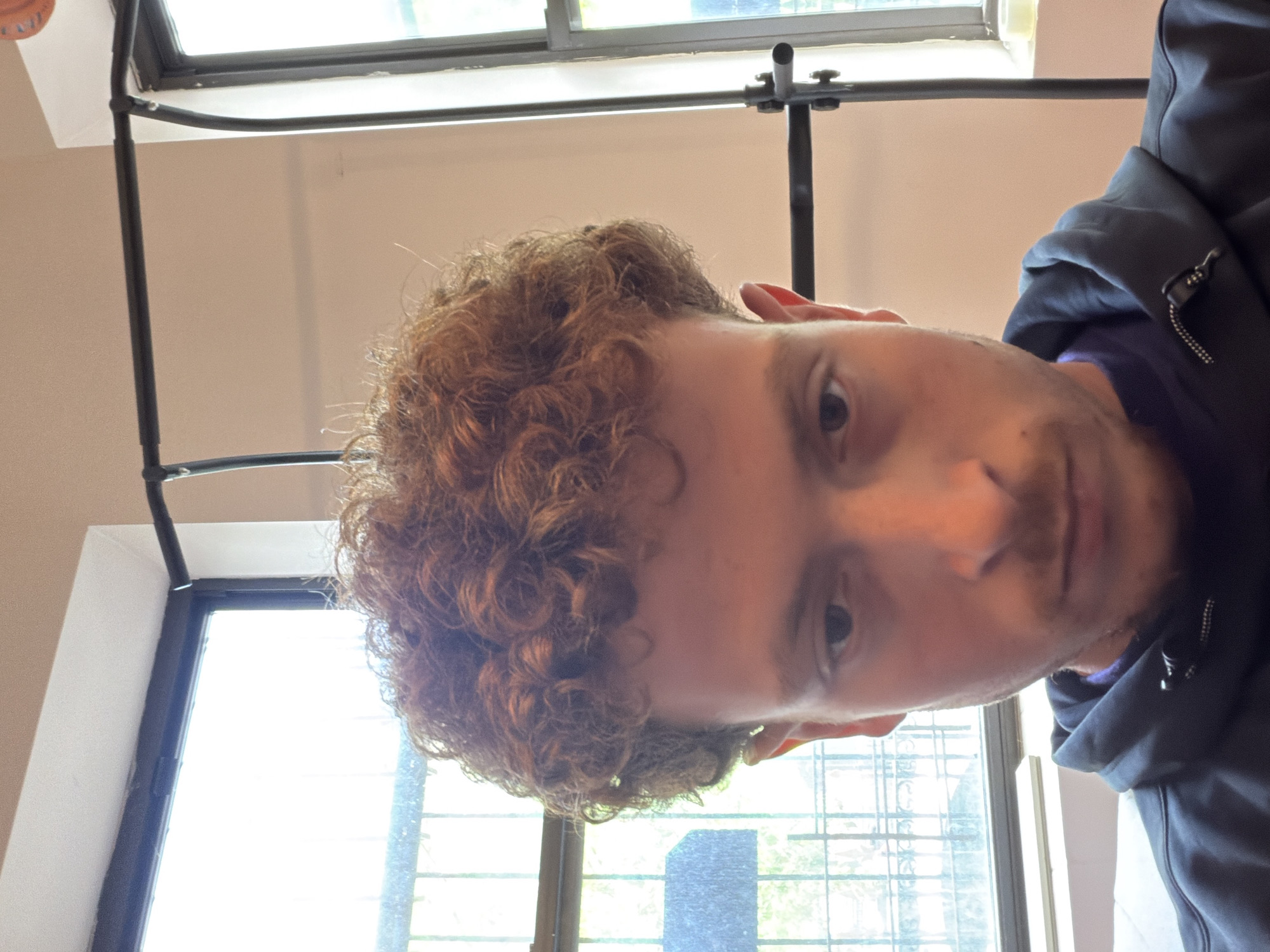 A selfie of a person with light skin and curly brown hair, wearing a dark hooded jacket. They are positioned indoors with windows in the background. The location is unidentified and it is not possible to confirm that it is New York City or even the United States.