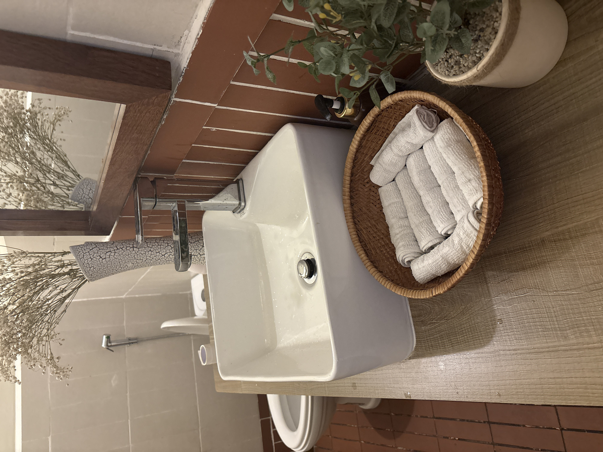 The image presents an eye-level, close-up view of a bathroom sink and countertop setup. The sink is a modern, rectangular white basin with a chrome faucet and drain. Beside the sink sits a round, woven basket filled with neatly rolled white towels. To the right of the towel basket is a potted plant featuring green foliage with gravel at the base, all sitting atop a countertop featuring wood grain. The wall behind the counter has a section of brown brick tiles, while the wall to the left of the sink and toilet are lined with neutral-colored square tiles. Visible in the background is a mirror, reflecting a vase of small white flowers. The toilet and shower are also visible, though partially obscured. Considering the provided location context, the photograph likely depicts a bathroom interior in Nha Trang, Vietnam.