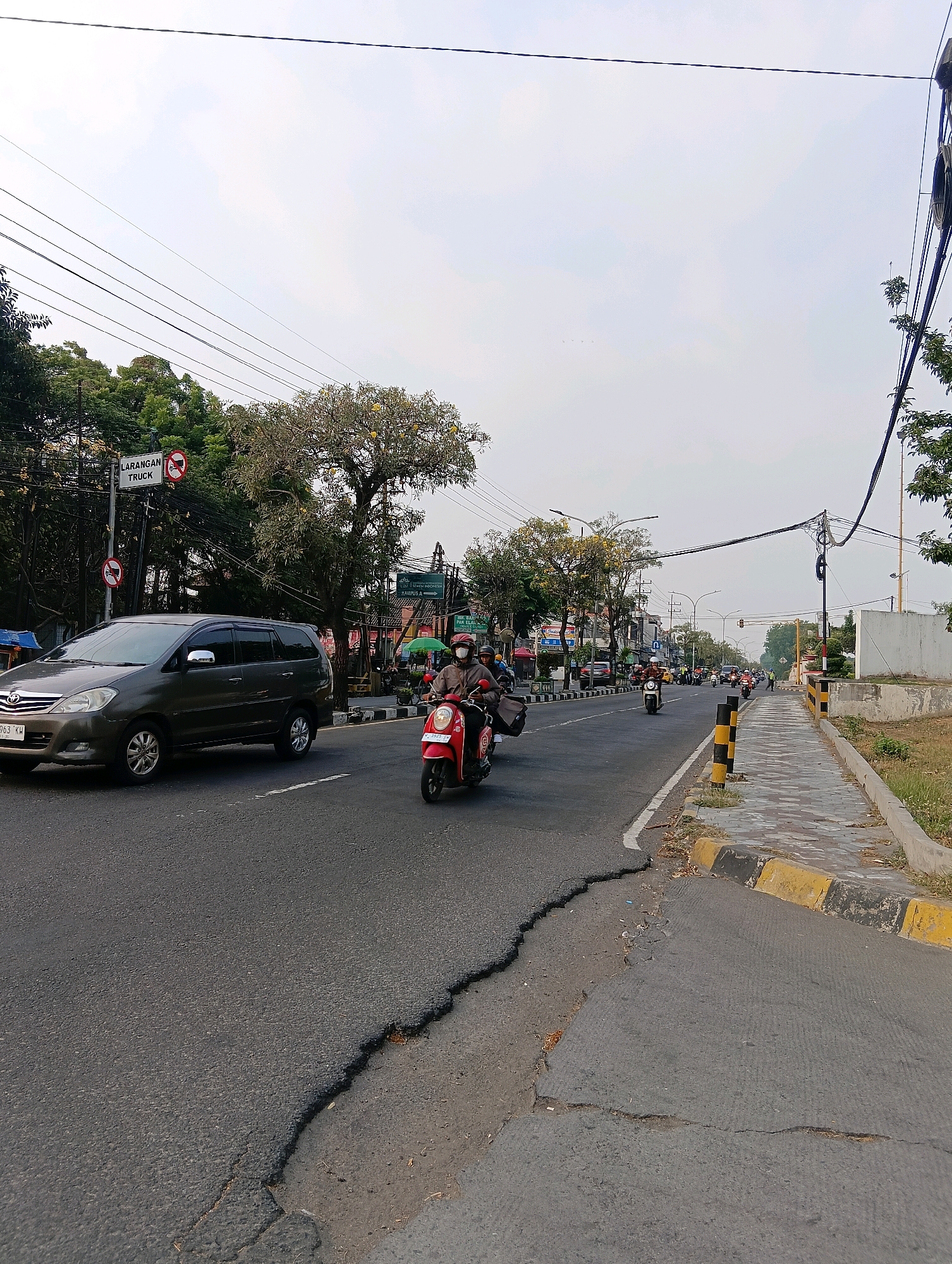The image depicts a street scene in Gapurosukolilo, Indonesia. A grey minivan is driving on the left side of the road, while a red scooter carrying two individuals, one driving and one passenger, is in the right lane. Both individuals on the scooter are wearing masks. The road has a cracked and uneven surface, particularly along the right edge bordering a paved sidewalk. The sidewalk is marked with yellow and black bollards. Trees line the street on both sides, and overhead power lines crisscross the sky. A sign on the left indicates "LARANGAN TRUCK," meaning "TRUCK PROHIBITED." The sky is overcast.