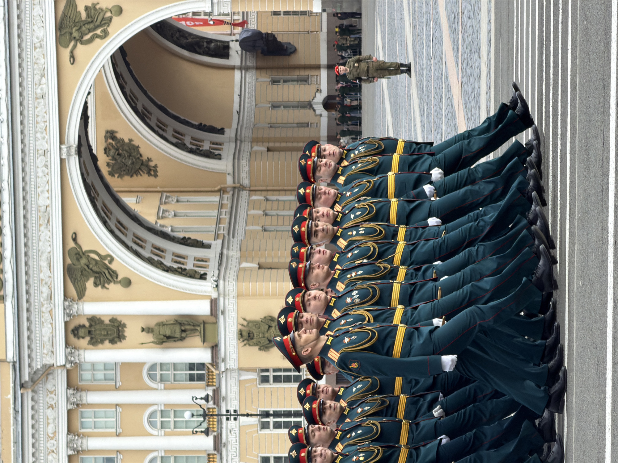 In Saint Petersburg, Russia, a military parade is in progress. Rows of soldiers in green uniforms with gold trim and red accents are marching in formation. They wear red caps and white gloves. The soldiers are passing under an archway of a building with ornate architectural details, including statues and decorative moldings. In the background, more soldiers and civilians are visible. The ground is paved with grey stones and lined with white stripes.