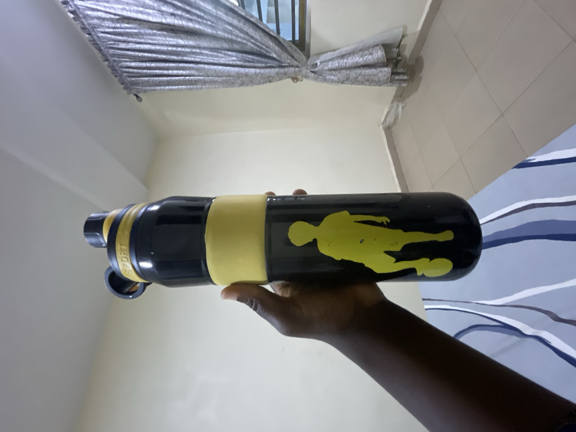 A hand with dark skin holds a black and yellow sports water bottle featuring a yellow silhouette of a person playing soccer. The bottle has "SPORT" written in yellow on its cap. The background consists of a light-colored wall, a window with grey curtains, and a tiled floor. The image appears to be taken indoors in a room. There is no visible indication of the specific location in Howayi, Nigeria.