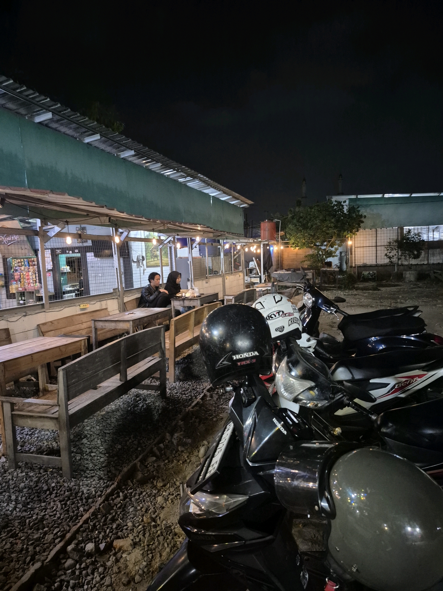The image shows an outdoor seating area of a small restaurant or warung in Pabelan, Indonesia, at night. Several wooden tables and benches are arranged on a gravel and dirt ground. A green building with a corrugated metal roof forms the backdrop, and strings of lights are visible along the eaves, illuminating the area.
In the foreground, a row of parked motorcycles is visible, some with helmets on their seats or handlebars. One motorcycle in the foreground has a black helmet with "HONDA TRX-1" written on it. Another motorcycle has a white helmet with "KYT" and "CRASH" written on it.
At a table in the middle ground, two individuals are seated. A man wearing a dark jacket is on the left, and a woman wearing a dark hijab is on the right. They appear to be conversing or eating. Behind them, a display of various items, possibly snacks or drinks, is visible on shelves inside the establishment. The overall atmosphere is casual and typical of a local eatery in Indonesia.