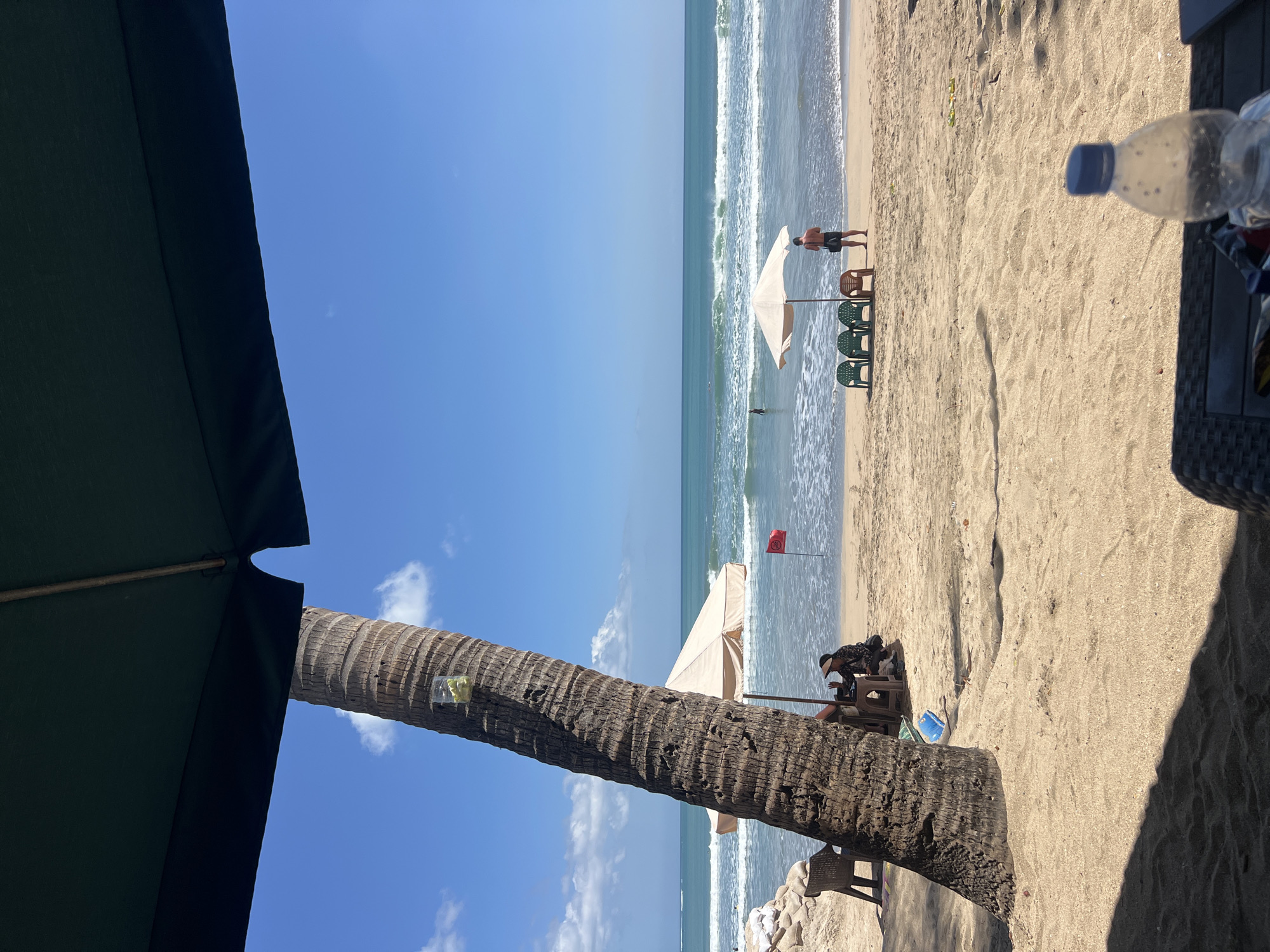 The image depicts a serene beach scene in Kuta, Indonesia, under a clear blue sky with scattered white clouds. In the foreground, a large palm tree trunk dominates the left side of the frame, its rough bark textured and weathered. Attached to the tree is a clear plastic bag containing some yellow items, possibly fruit. Partially visible in the foreground are shadows cast by an overhead umbrella, and a table with a plastic water bottle and other items on it.
The sandy beach stretches towards the ocean, where gentle waves are rolling in. Several beach chairs are arranged on the sand, some unoccupied and others with people seated under a cream-colored umbrella. A person in black shorts stands near the umbrella, facing the sea. Further out in the water, another individual is visible. To the left, there's a structure made of sandbags, and another beach chair. A small red flag planted in the sand near the water's edge signifies a safety marker. The overall atmosphere is one of a tranquil day at the beach, with a hint of relaxation and leisure.