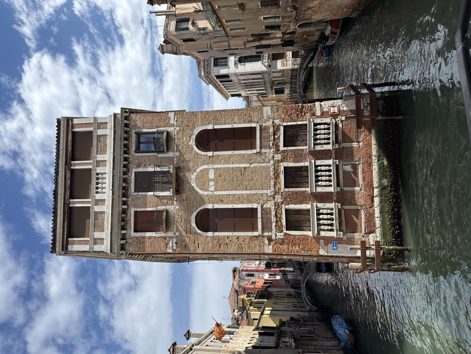 The image depicts a canal junction in Venice, Italy, likely at the confluence of the Rio del Santissimo and Rio de la Chiesa, near Fondamenta Zattere.
In the foreground, a prominent five-story, weathered reddish-brown brick building rises directly from the dark green canal water. Its lower visible floors feature windows with white stone balustrade balconies and closed brown wooden shutters. The central window openings on the first and second visible floors are bricked-up, retaining their arched outlines. The third floor has windows with minimal iron railings, and the top floor displays three windows with closed brown roller shutters, set behind a white ornamental cornice. A blue arrow sign pointing left and a red "2.0" marker are affixed to the lower left base of the building, where wooden mooring poles are also visible.
Two canals extend from the central building. The left canal features a blue-tarped boat moored by a multi-story building line, where a Venetian flag is visible on a pole. A stone arch bridge, traversed by several indistinct human figures, spans this canal in the mid-distance. The right canal also shows several small, dark boats moored along its bank, with another stone arch bridge visible further along its course.
The surrounding environment consists of dense, multi-story Venetian buildings with varied facades (yellow, red, white, beige) and terracotta roofs, characteristic of the city's architecture. The sky is partly cloudy, displaying patches of blue interspersed with scattered white cumulus clouds. The visible human figures are walking on the distant bridge.