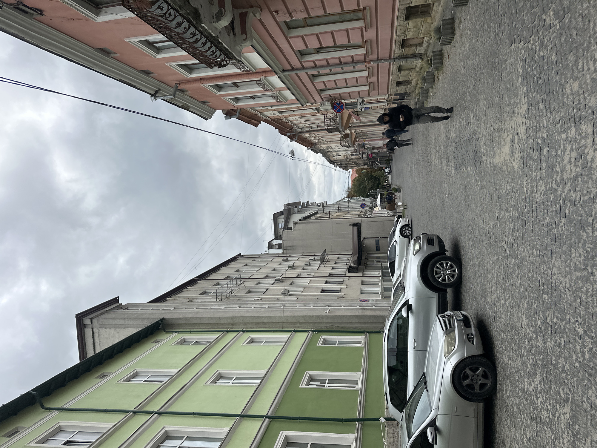 The image depicts a street scene in Ternopil, Ukraine, likely on a cloudy day. On the left side of the street, there is a bright green building with multiple windows and white trim. Adjacent to it is a taller, grey building with a more utilitarian design, featuring numerous windows and external air conditioning units. On the right side of the street, a salmon-pink building with decorative balconies and classical architectural elements lines the sidewalk. The street itself is paved with cobblestones. Several cars are parked along the left side of the street, including a silver sedan in the foreground and a silver SUV behind it. Further down the street, several people are walking. One man in the foreground is wearing a black hooded jacket and is looking at his phone. The sky is overcast, suggesting a potentially cool or damp day. The overall impression is of an urban environment with a mix of architectural styles.