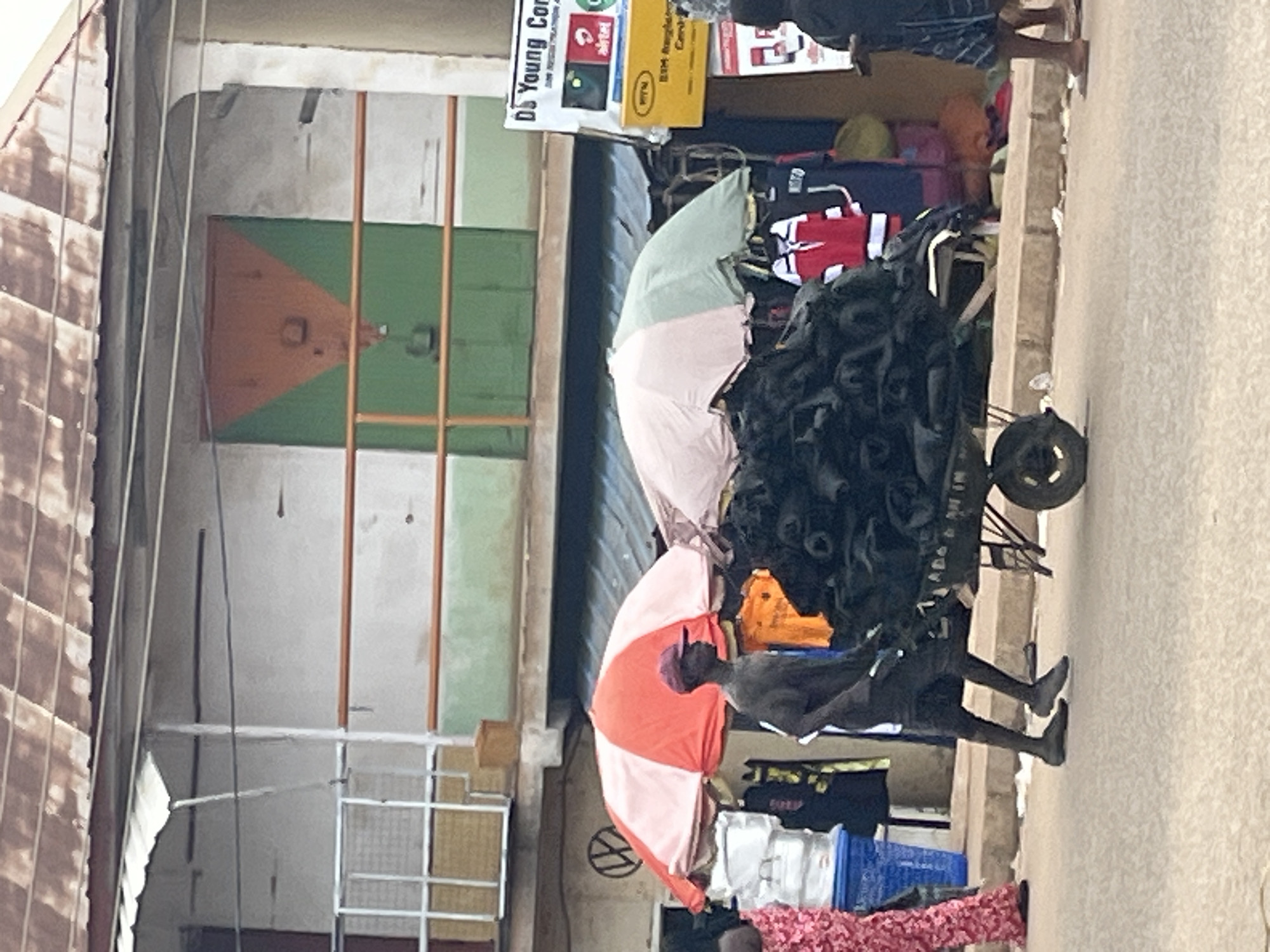 In Bukuru, Nigeria, a man wearing a dark shirt and shorts, and a maroon cap, walks beside a cart loaded with black tires. The cart is being pushed from the left to the right. Behind the man, market stalls are visible with colorful umbrellas, including a red and white one and a pink and green one. Several people are visible in the background, including a person in a pink patterned dress on the left, and another person wearing a dark shirt and plaid shorts on the right. A white building with a rusty corrugated roof is situated behind the market stalls. Signs are posted on the right side of the building, one of which appears to be for "DS Young Com." The scene depicts a busy street market environment.