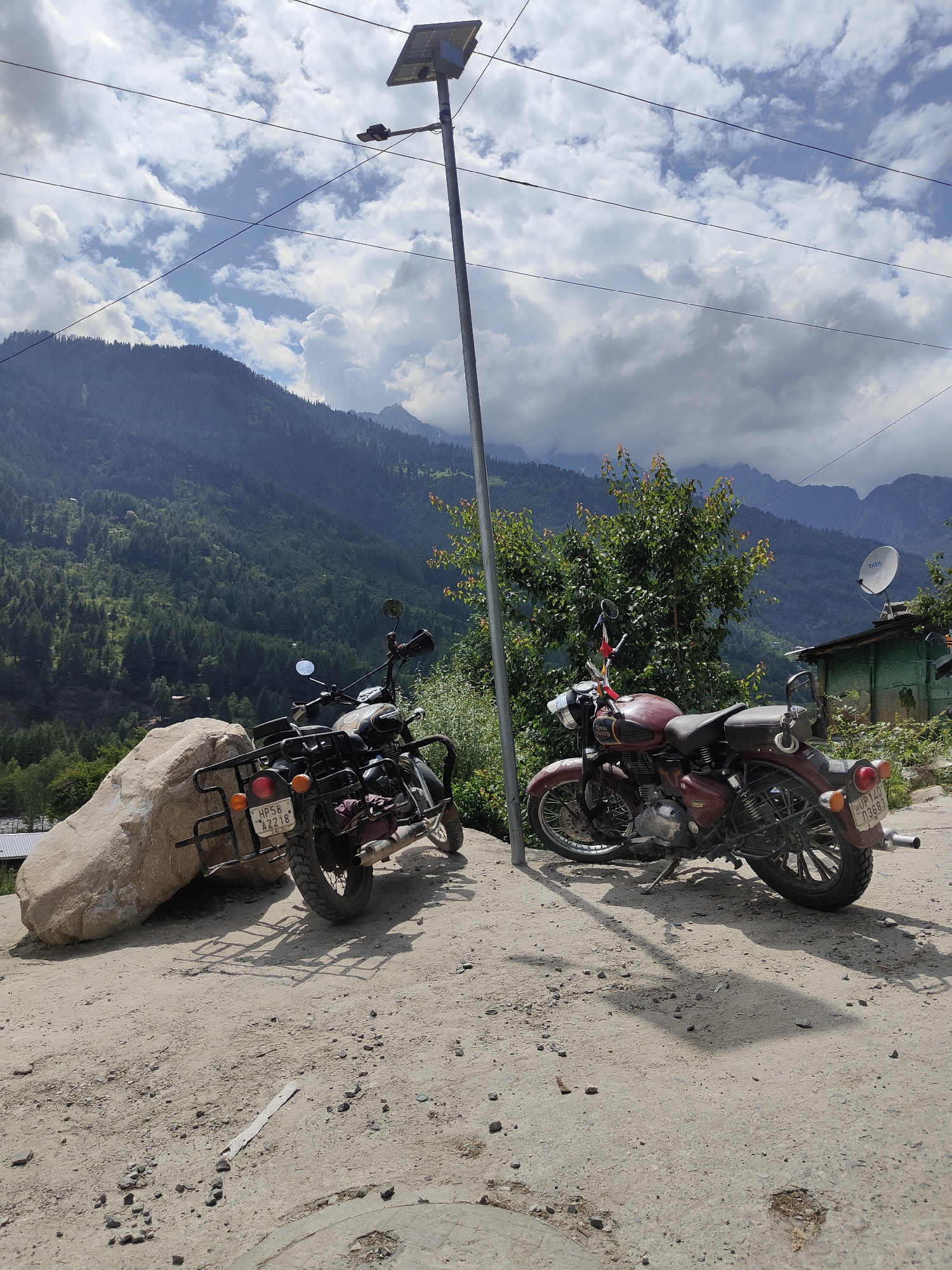 The image captures a scenic view in the mountainous region of Vashisht, India, under a partly cloudy sky. In the foreground, two motorcycles are parked on a dirt road. The motorcycle on the left is dark colored with a license plate that reads "HP58 A2218". It is positioned next to a large, light-colored boulder. The motorcycle on the right is a maroon Royal Enfield with a license plate that reads "UP 14D D3683". It is parked under a solar-powered street lamp.
Behind the motorcycles, a dense green forest covers the steep slopes of a mountain, reaching up towards the sky. The mountains are layered, with some peaks visible in the distance. A small, green building with a satellite dish on its roof is visible on the right side of the image. The overall scene suggests a remote and picturesque location, likely a stop for travelers exploring the natural beauty of the area.