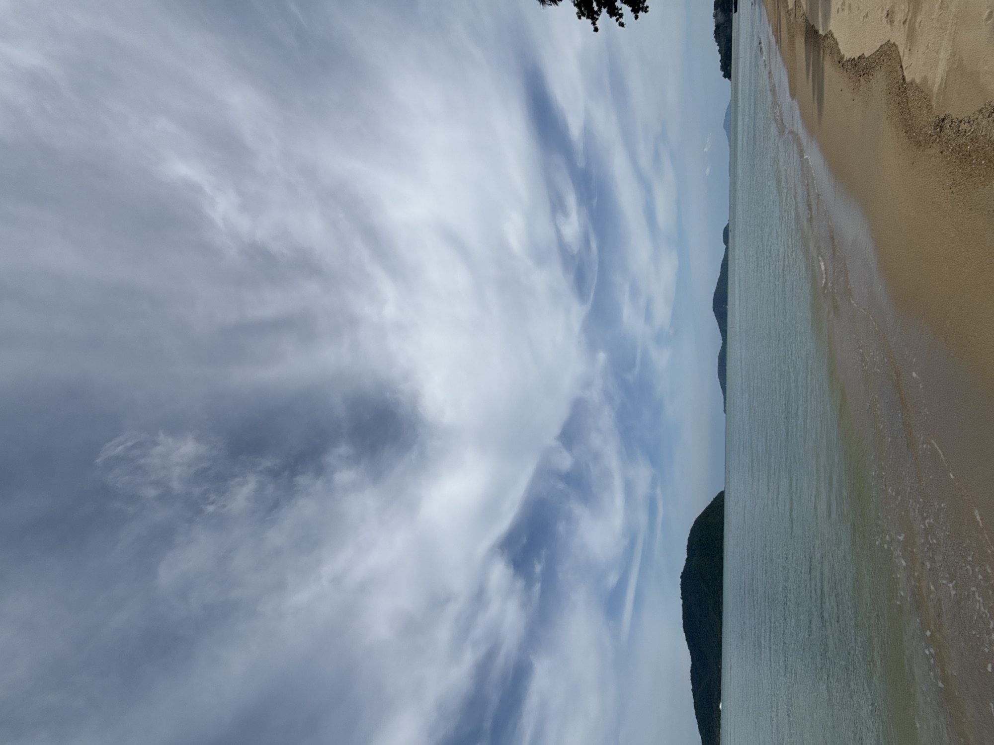 An aerial view of a tropical beach in Rawai, Thailand, features a serene landscape with a sandy shore meeting the calm, turquoise waters of the Andaman Sea. Lush green islands dot the horizon, some with steep hills and dense vegetation. The sky is a canvas of dramatic, wispy clouds, with streaks of white and grey against a pale blue backdrop, suggesting a gentle breeze. The foreground showcases the soft, golden sand of the beach, with gentle waves lapping at the shoreline, leaving behind a delicate foam. The overall scene evokes a sense of peace and natural beauty, characteristic of a secluded tropical paradise.