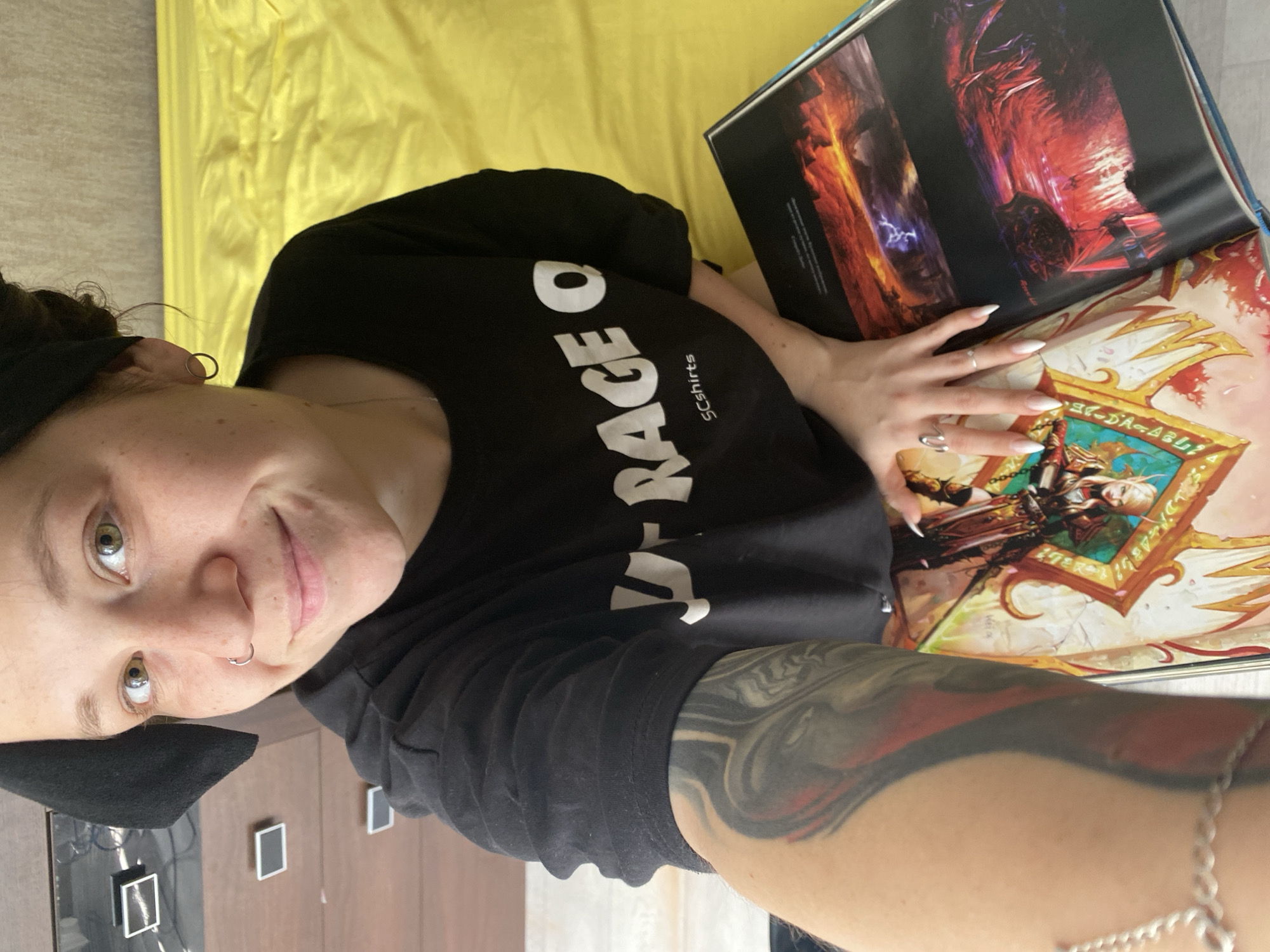 This is a vertically oriented selfie captured from a high angle, featuring a young person lying down and engaging with a large art book.
The main subject is a young person, likely female, with dark hair pulled back with a black headband or scarf. They have light-colored eyes, a septum piercing, and a subtle, relaxed smile directed at the camera. Their left hand, adorned with two silver rings and long, pointed white manicured nails, rests gently on the open book. A prominent, colorful tattoo, primarily in black, grey, and red tones, covers their right forearm, which is also embellished with a silver chain bracelet. They are wearing a black t-shirt with white text, partially visible as "RACE O" and "SCshirts".
The person is holding a large, open art book displaying vivid fantasy illustrations. The left page shows a dramatic scene with dark, fiery orange and purple elements, including a striking lightning bolt. The right page features an intricately framed illustration of an elven-like warrior character, equipped with a bow, chainmail, and a sword, set against a background of white and gold. Partial text "GC-DA-ABL" is visible near the bottom of this illustration.
The setting appears to be an indoor space, likely a bedroom or living area. The person is reclining on a bright yellow fabric surface, possibly a bedspread or blanket. To the upper left in the background, a wooden surface is visible, featuring two modern black rectangular elements, potentially outlets or controls, with some wires.
The activity taking place is a relaxed moment of reading or admiring art, combined with taking a selfie. The person's expression conveys a sense of calm and contentment. While the image itself lacks specific geographical indicators, the provided context places this scene in Chaikovsky, Russia.