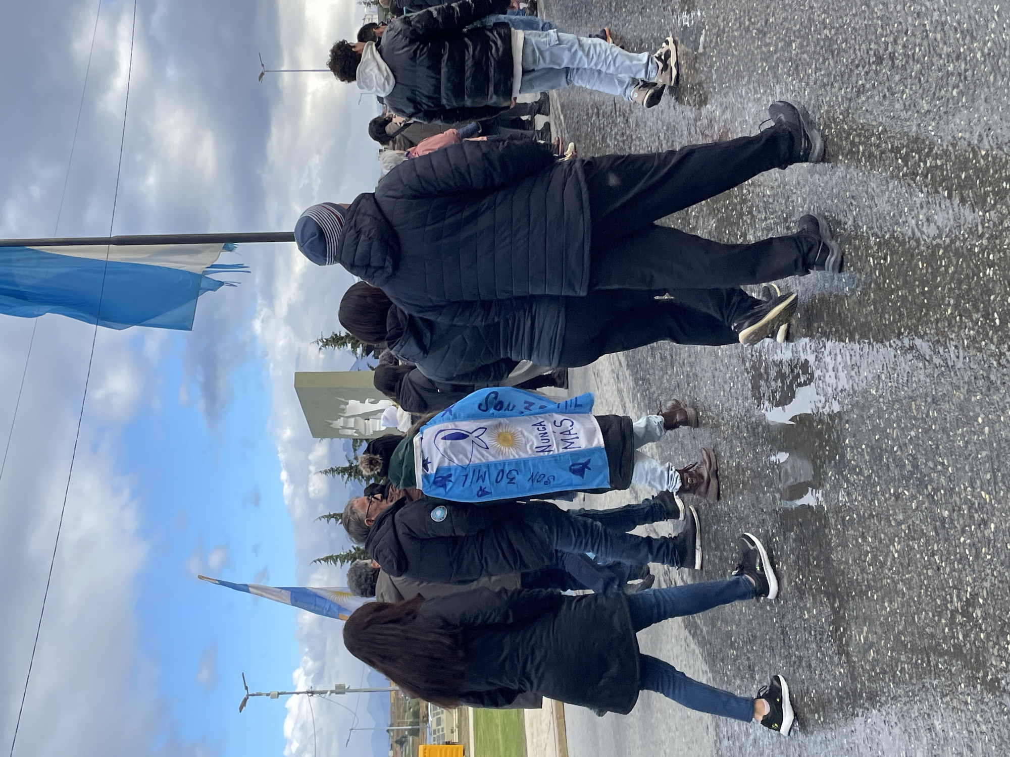 The image depicts a group of approximately 10 to 15 individuals walking on a wet, paved street surface in Ushuaia, Argentina. The sky above is partly cloudy with patches of blue.
The street shows visible puddles and reflections. Most pedestrians are dressed in dark, cold-weather clothing, including puffer jackets, long coats, and jeans. One prominent individual, viewed from behind, wears a blue and white poncho-style jacket designed to resemble the Argentinian flag, bearing the white symbol of a headscarf (Mothers of the Plaza de Mayo) and text that reads "Son 30 MIL" and "NUNCA MAS."
A large, light-blue and white Argentinian flag, visibly frayed or torn at its lower edge, flies on a dark pole on the right side of the frame. Another smaller Argentinian flag is visible on a pole further down the street on the left. Behind the pedestrians on the right, a tall, angular, light-colored monument or memorial with an irregular etched design is partially visible. Coniferous trees line the street behind the monument, and distant mountains are discernible in the background beneath the cloudy sky. A streetlight pole is visible on the left side. The individuals are uniformly walking in the same general direction away from the viewer. No direct interactions between individuals are discernible.