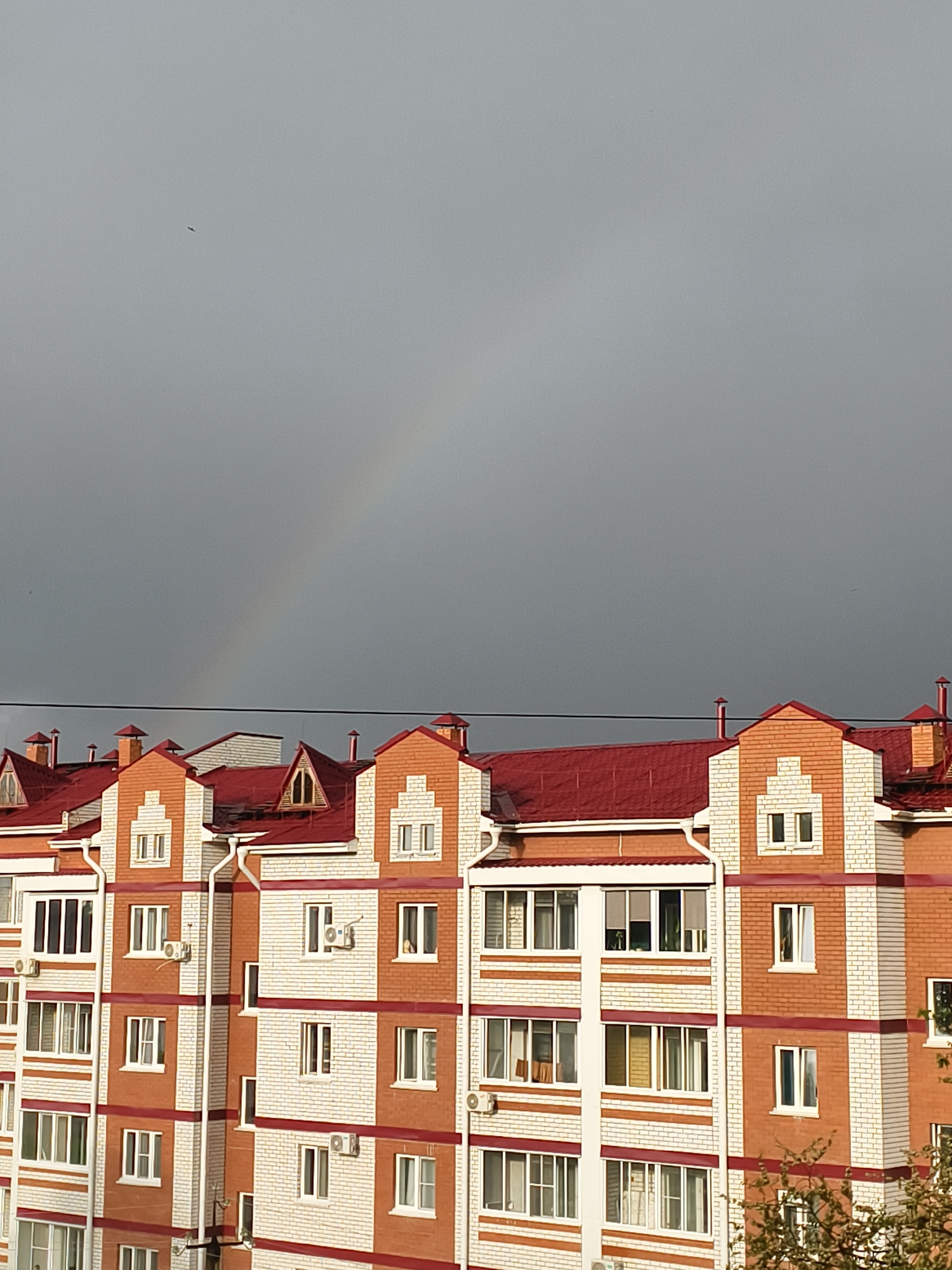 The image displays the upper portion of a multi-story residential building set against a dark, overcast sky. The building features a striking design with red-tiled roofs, prominent gables, and facades constructed from alternating patterns of reddish-brown and white or light-colored bricks. Numerous windows are visible across the different levels, some equipped with balconies or external air conditioning units.
In the upper half of the frame, the sky appears a uniform dark gray, suggesting heavy cloud cover or impending rain. A faint and subtle rainbow arc is discernible in the middle-right section of the sky, indicating the presence of sunlight breaking through moisture in the atmosphere, likely after or during light rain. A small, dark, indistinct flying object, possibly a bird or insect, is visible in the upper-left quadrant of the sky.
A thin, dark power line stretches horizontally across the image, positioned just above the rooftops of the building in the center-right. The building's architecture includes multiple roof levels and dormer windows, adding to its complex profile. The illumination on the building's facade, despite the dark sky, suggests it is daytime, with light casting some shadows and highlighting textures. The scene depicts an urban or suburban residential area in Belogorsk, Russia. There is no visible text in the image.