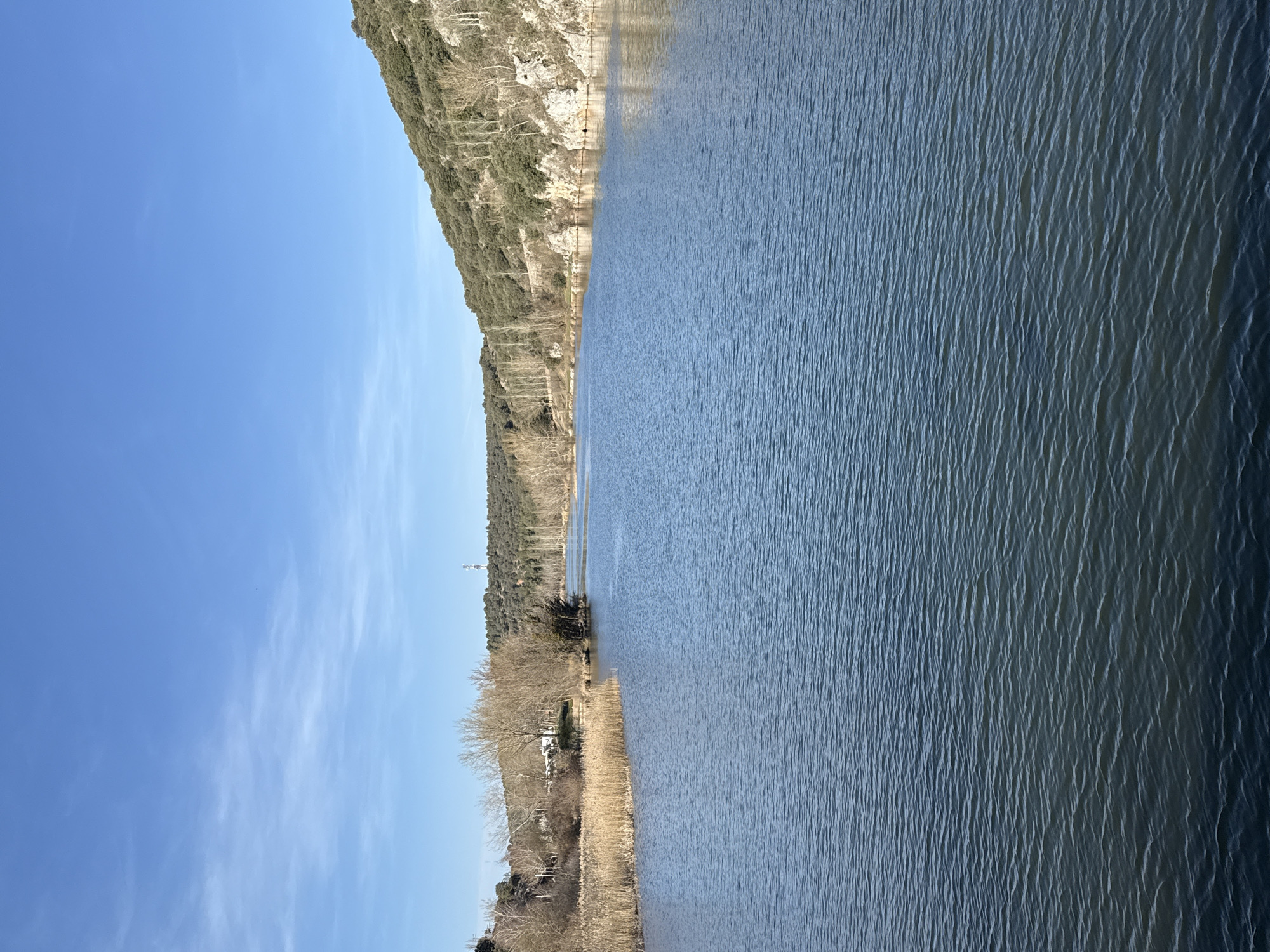 The image depicts a wide view of the Duero River in Soria, Spain, under a clear, bright blue sky with sparse, wispy white clouds. The river occupies the central and lower portions of the frame, displaying a dark blue-grey surface with numerous small ripples. On the left bank, a dense expanse of dry, golden-brown reeds extends along the water's edge in the midground. Behind these reeds, a cluster of bare deciduous trees with thin branches is visible, followed by a gently sloping hillside covered in similar dormant vegetation. A white, oblong structure, possibly a recreational vehicle or small building, is partially obscured among these trees. Further in the distance, a tall, slender telecommunications tower with red and white markings stands atop a distant hill. The right bank features a steeper, rugged hillside characterized by exposed light-colored rock formations and covered with dark green evergreen shrubs interspersed with bare deciduous trees. The river flows from the foreground towards the background, where it appears to narrow slightly between the receding banks. No individuals, animals, or watercraft are present in the scene.
