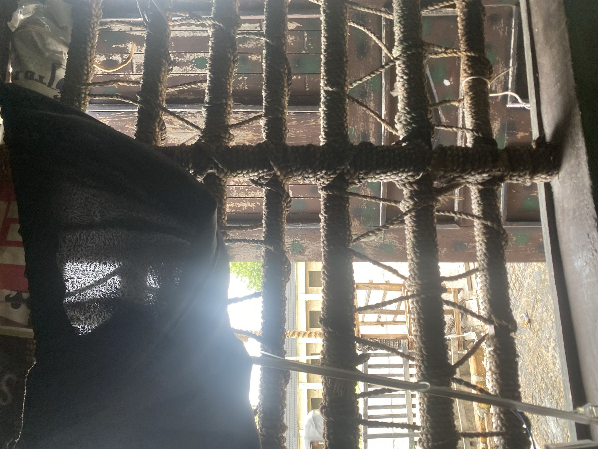 This media file captures a close-up view of thick ropes, likely part of a makeshift barrier or structure, strung vertically and horizontally. Behind the ropes, a weathered wooden surface with peeling paint is visible, hinting at an outdoor or aged setting. To the left, a dark, draped fabric obscures a portion of the scene, with some indistinct patterns or text on a lighter material peeking out from behind it. Sunlight streams in from the top left, creating a bright lens flare and illuminating some of the rope textures.
Through the gaps in the ropes, glimpses of a different environment can be seen. There appears to be another structure with wooden beams and what might be a ladder or scaffolding. Further in the background, hints of open space and possibly trees suggest an outdoor location. A small, indistinct figure in white can be seen in the distance. The overall impression is one of a somewhat rustic, possibly unfinished, or utilitarian space. The lighting suggests it is daytime. The location context is Jalingo, Nigeria.