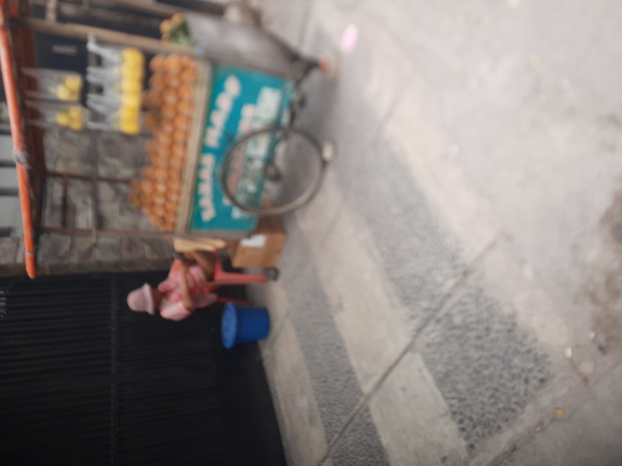 In the city of Central Jakarta, Indonesia, a blurred image captures a street scene. To the left, a person wearing a pink hat and a pink patterned shirt sits on a red chair, leaning against a dark fence. In front of them, a blue plastic bucket sits on the ground. To the right, a food cart is visible, filled with orange-colored fruits, possibly oranges or tangerines. Above the fruit, several plastic bags hang, each containing yellow items, likely more fruit. The cart has a turquoise sign with white lettering that is partially obscured. A bicycle wheel is attached to the cart. In the background, a stone wall and a wooden structure are present. The ground is paved with concrete tiles, some of which are patterned with dark grey pebbles.