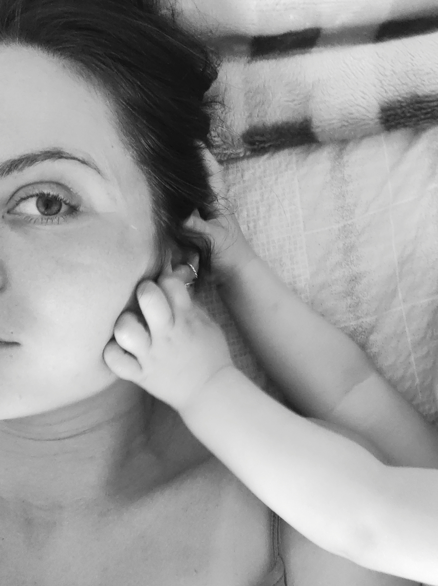 A close-up, black and white photograph depicts the face of a woman, her left eye visible and looking directly at the camera. A small child's hand is touching the woman's face near her ear, the child's arm extending towards the right side of the image. The background appears to be a textured surface, possibly a blanket. The location is unknown.