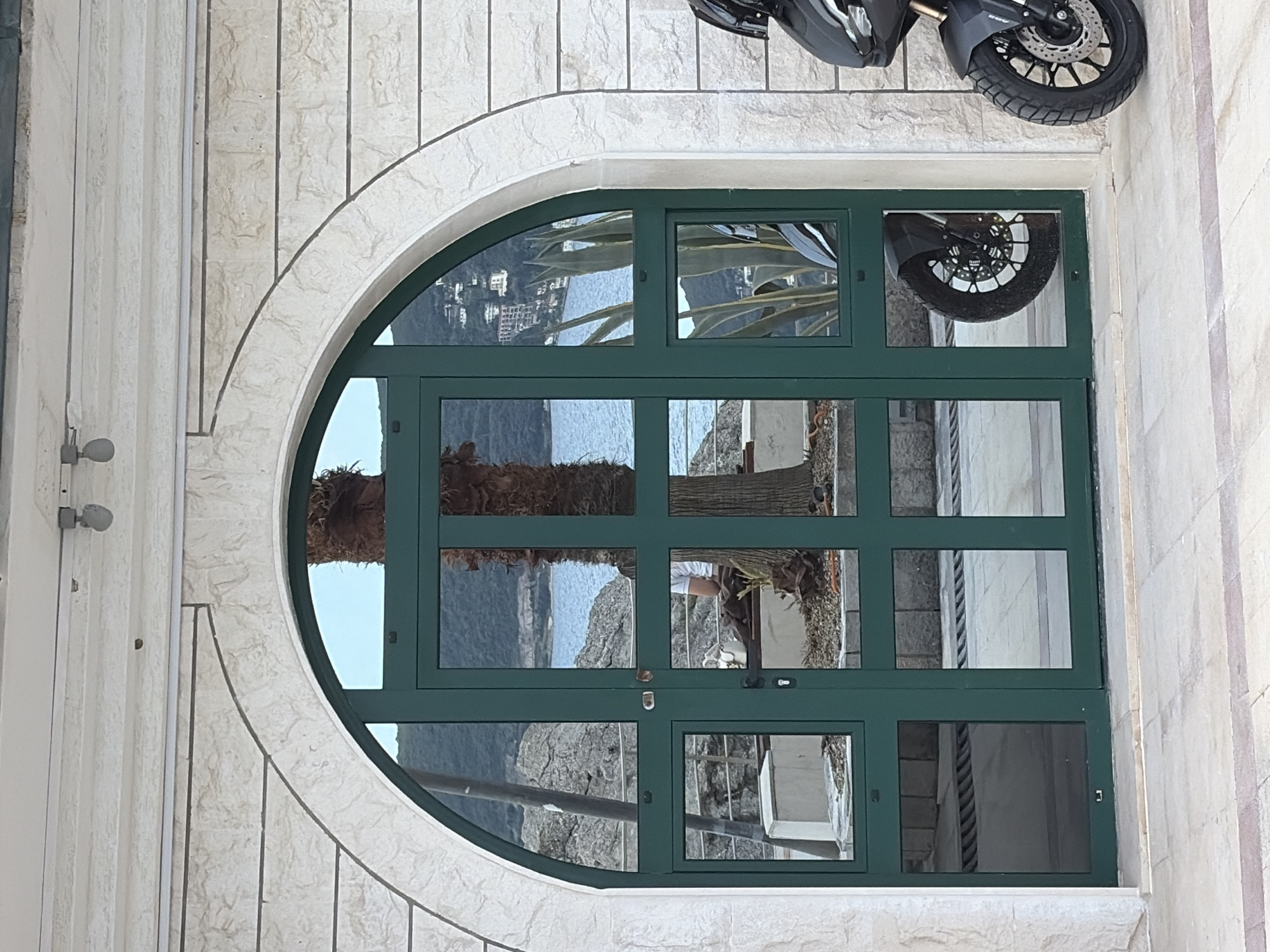 A horizontal outdoor shot features a building in Herceg Novi, Montenegro. The building’s wall is white with vertical lines. A semicircular arched doorway is the primary focus. The door is green with multiple rectangular panes of glass. The glass reflects the exterior environment, including a body of water, trees, buildings, and a portion of a motorcycle. The reflection also captures a person. The upper right corner of the frame shows part of the motorcycle parked near the doorway.