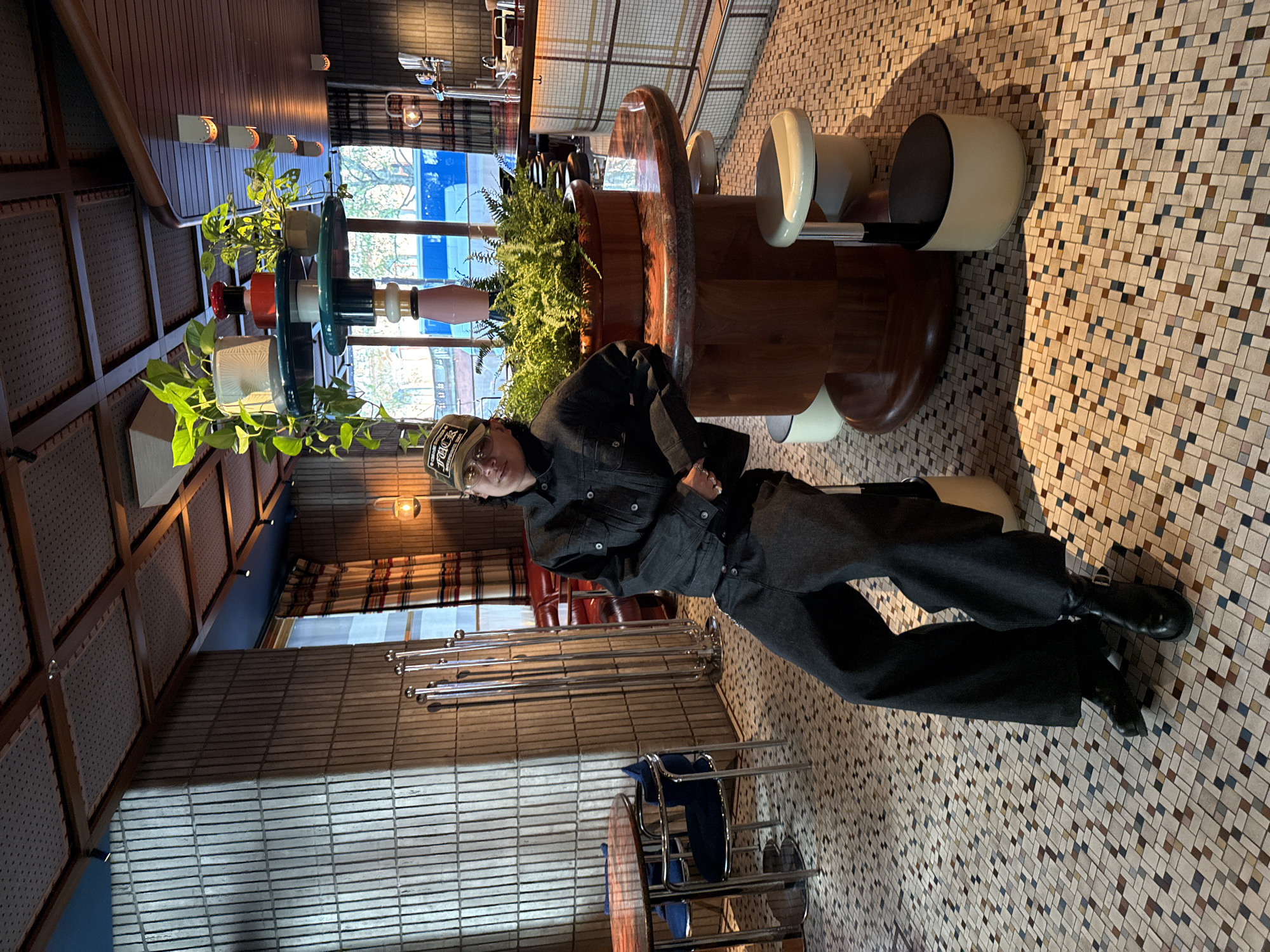 A person is captured posing in a stylish bar or cafe interior. The individual, dressed in a dark denim jacket, dark wide-legged trousers, and dark boots, is seated or leaning casually, looking towards the camera with a neutral expression. They wear a baseball cap with visible text, possibly "FYCK".
The setting features a distinctive multi-colored tiled floor, composed of small square tiles in various shades of beige, brown, and grey. A prominent, curved bar or counter, seemingly made of wood with a polished surface, anchors the space. Abundant green plants, including ferns and leafy potted plants, are arranged along the bar, interspersed with decorative items in red, blue, and white hues.
In the background, large windows reveal natural light and glimpses of trees and buildings outside, suggesting it is daytime. The interior walls exhibit varied textures, including horizontally striped panels and acoustic-like ceiling panels. Several bar stools with light-colored, circular seats are positioned at the counter. Other metal chairs and tables are visible further back in the venue. The overall atmosphere conveys a modern and inviting establishment.