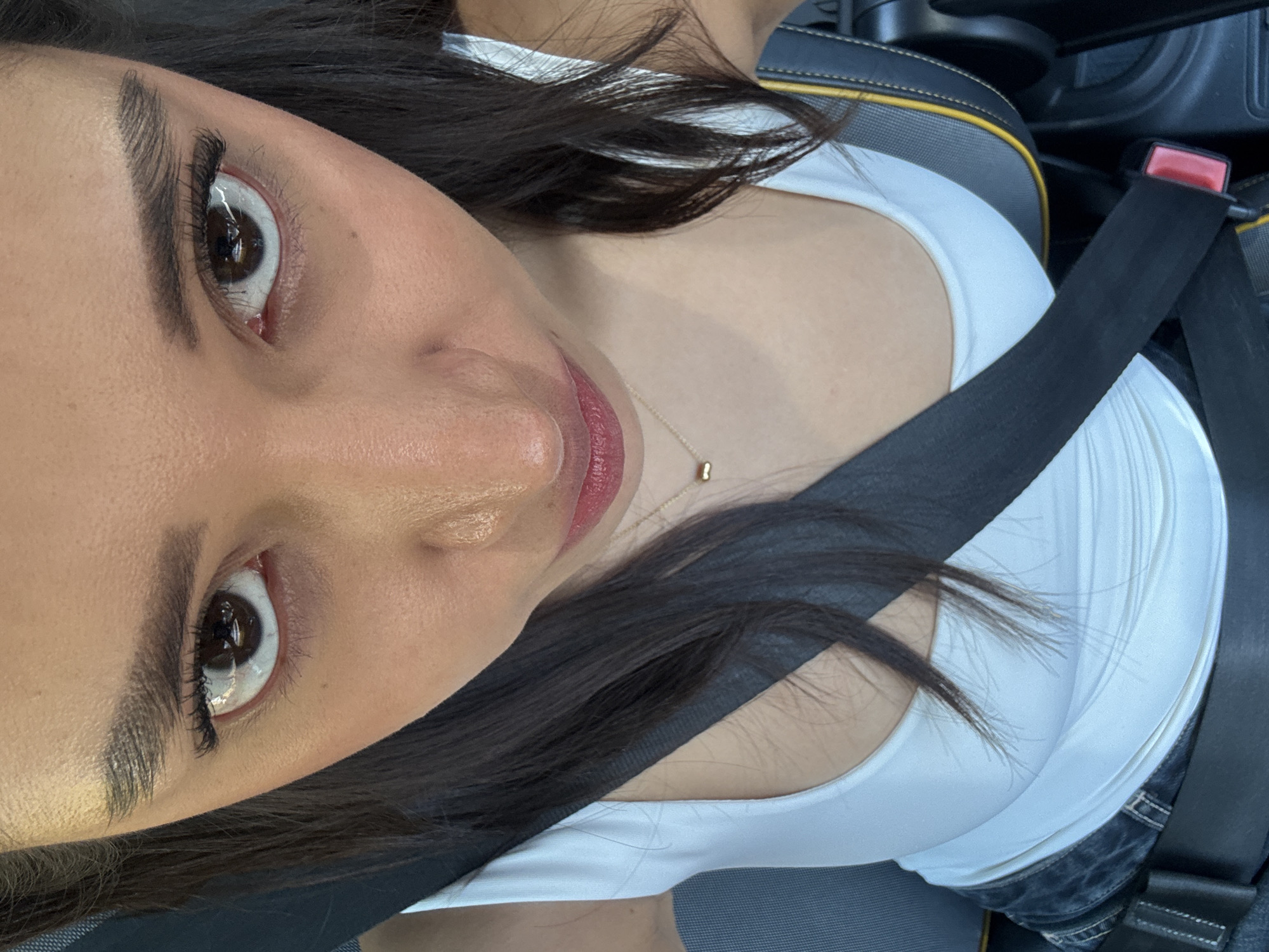 A selfie from a woman in a car, wearing a white tank top, a gold necklace, and a seatbelt. The location appears to be inside a vehicle, potentially in transit, but there is no additional information in the image to indicate that it is in Puebla, Mexico.
