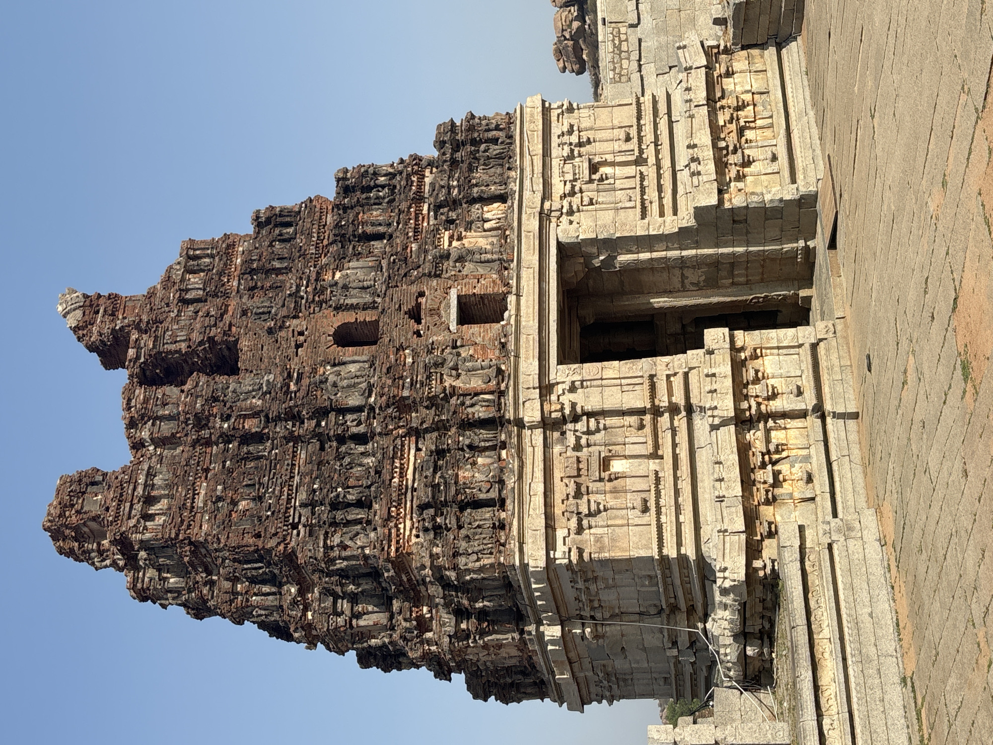 The image shows the Virupaksha Temple in Hampi, India. The temple's gopuram, or tower, is prominently featured, displaying intricate carvings and multiple tiers. The structure is partially constructed from a dark, weathered material, contrasting with the lighter stone of the base and surrounding areas. The gopuram’s layered design includes small openings or windows. The entrance shows detailed stonework and depth into the interior. The foreground includes stone paving. The sky above the temple is clear and blue.