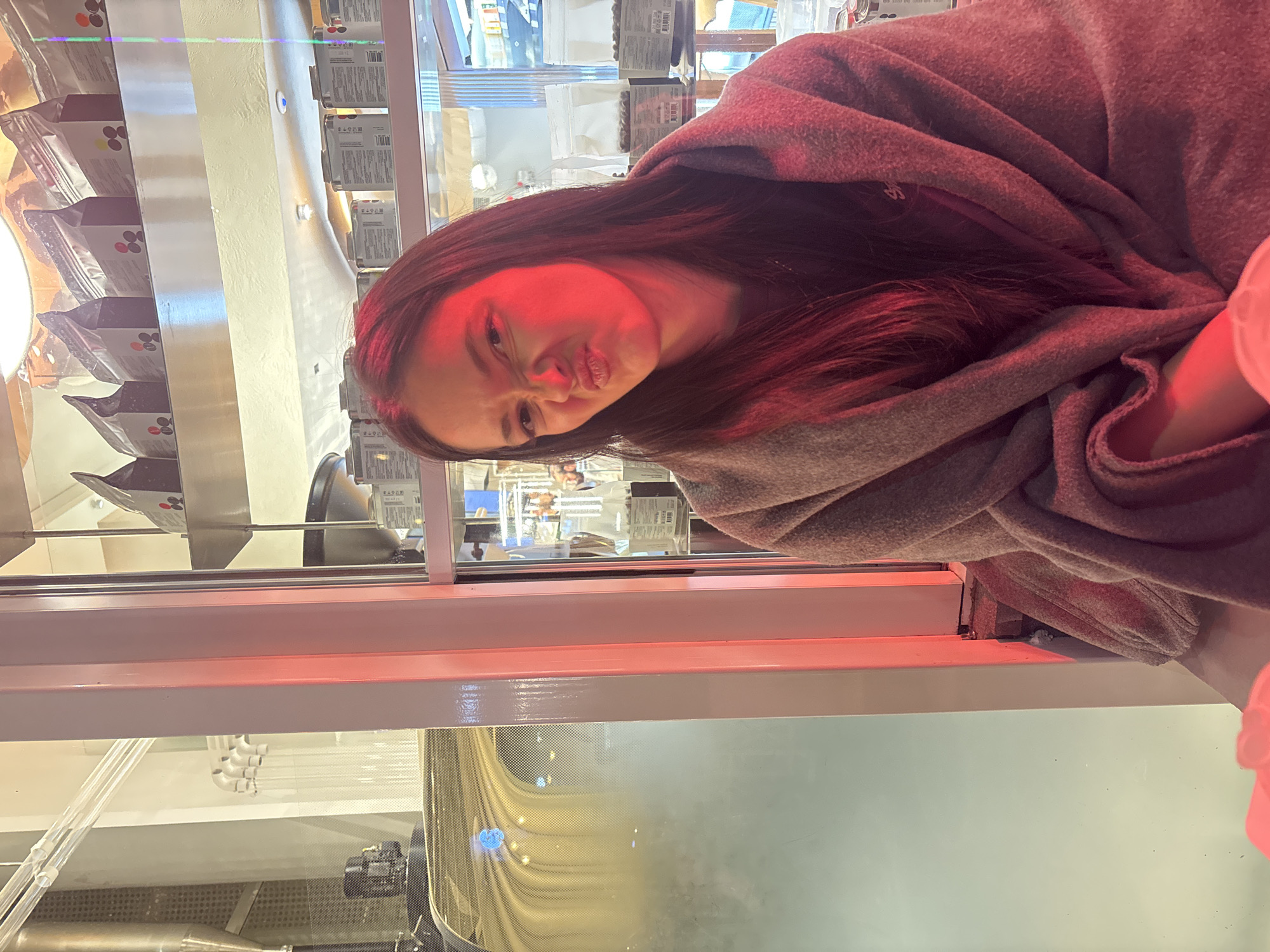 A woman with long dark hair is posing in front of a shop window. The shop has shelves stocked with packaged goods. The red tint over the woman suggests an indoor setting with specific lighting. There is no clear indication of Yerevan, Armenia, in the provided image details.