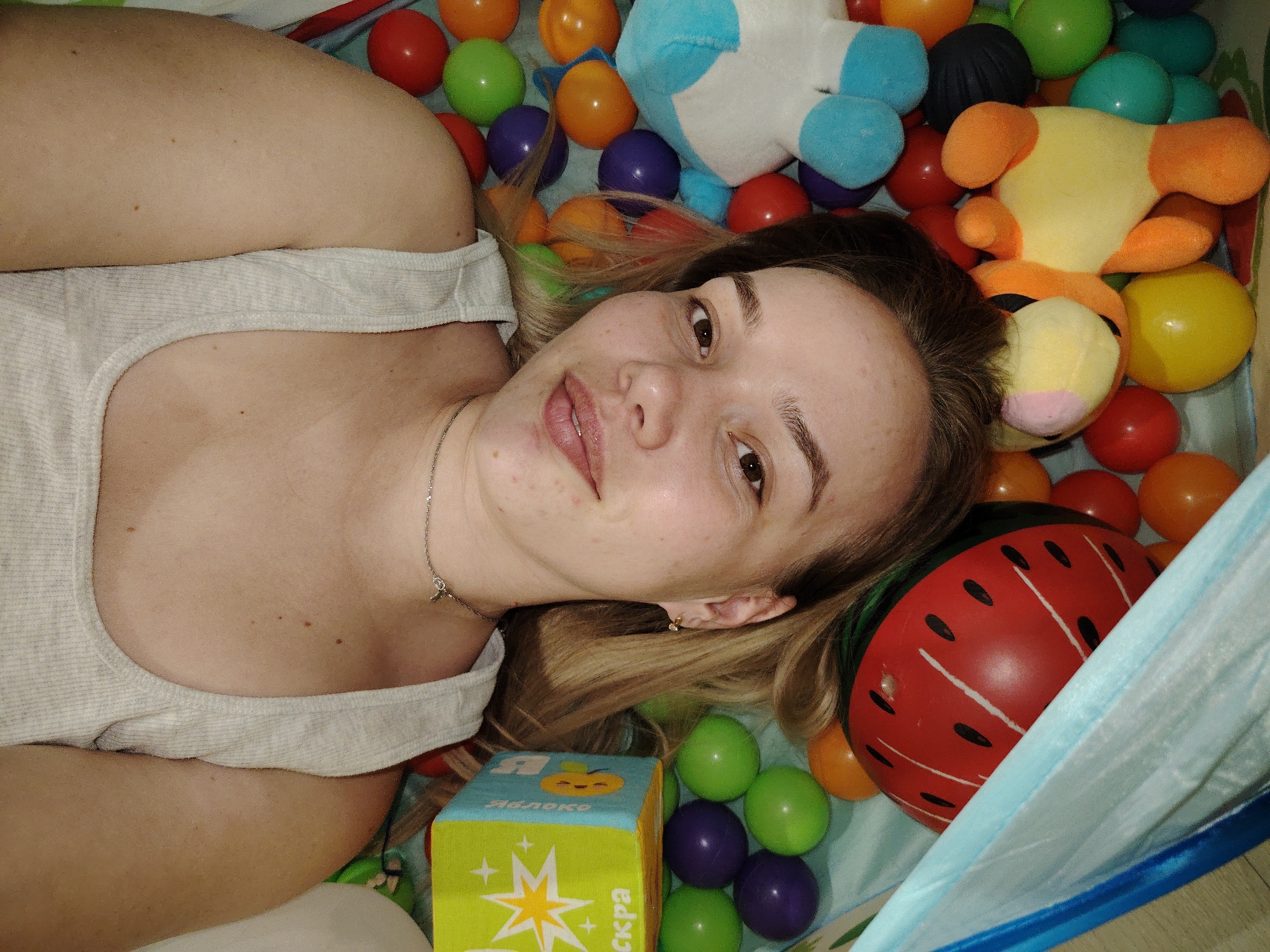 A woman is lying down in what appears to be a child's play area filled with colorful plastic balls and plush toys. The location is not specified, and there is no definitive information within the image to ascertain that the location is in Марковский, Russia. The woman has light skin and light brown hair, and she is wearing a light-colored tank top. She is looking directly at the camera with a slight smile. Around her, there are various colorful plastic balls and stuffed toys, including a blue and white plush unicorn and a yellow and orange plush animal. A green and yellow block with Cyrillic text is also visible. The surrounding environment is blue and white, possibly indicating the inside of a play tent or ball pit. A red helmet decorated as a watermelon is next to her head.