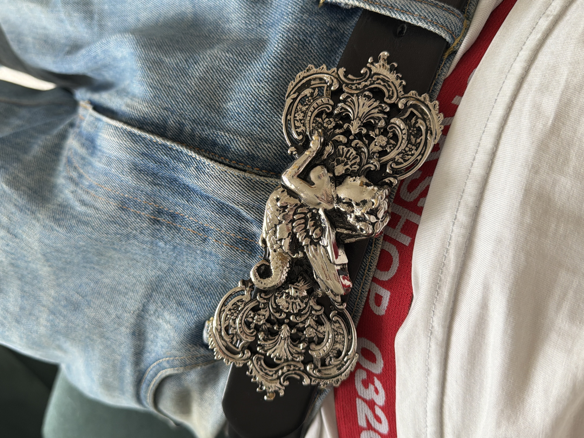 A close-up shot shows a person wearing light-wash denim jeans and a black leather belt with a large, ornate silver-colored buckle. The buckle features a human-like figure intertwined with decorative floral and swirling designs. Part of a white t-shirt with a red band that reads "SHOP 032C" is visible next to the belt. The environment is nondescript, with a focus on the clothing and accessories. The location is not discernible from the visual details provided.