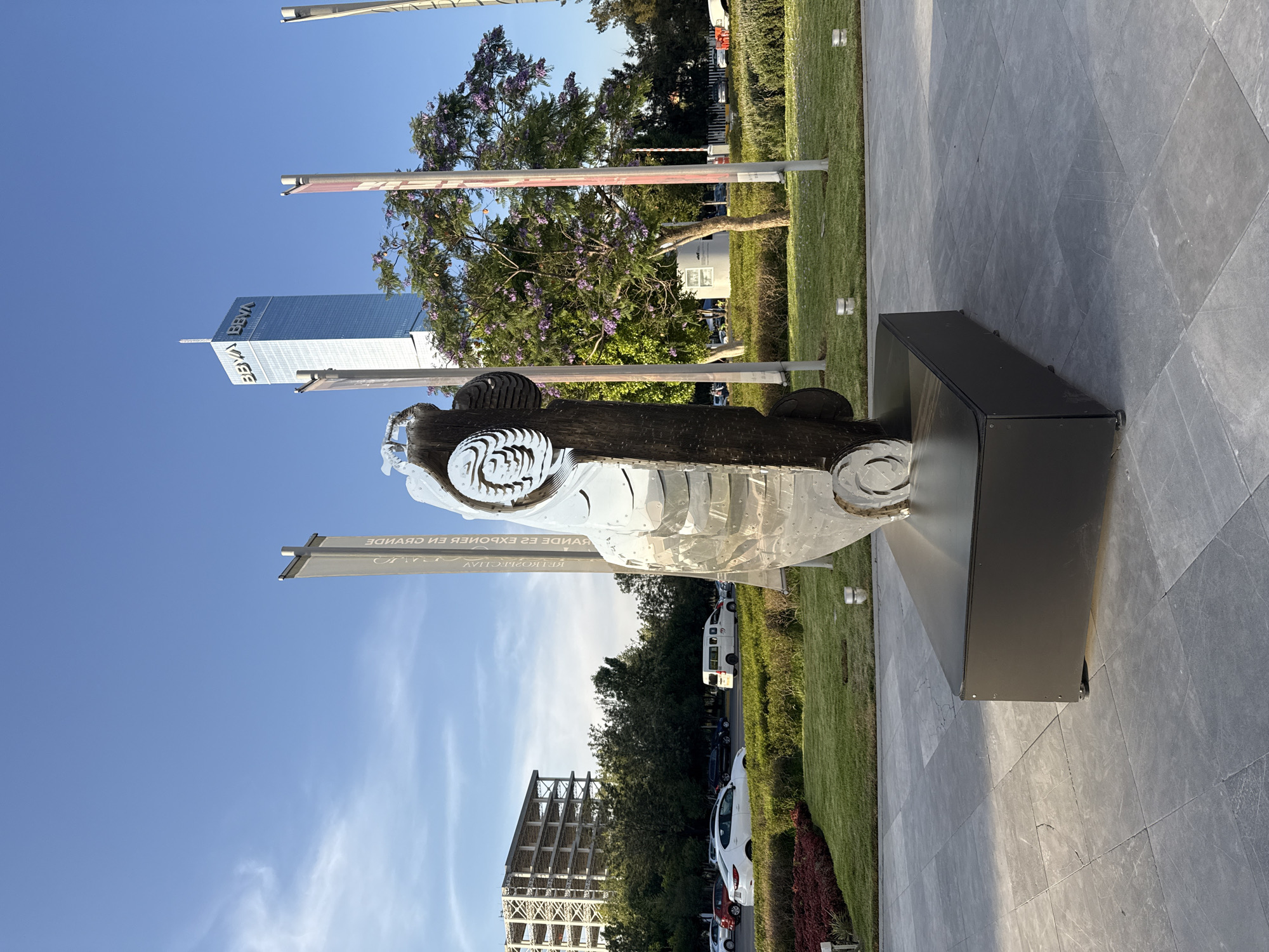 A sculpture of a car sits on a black rectangular base on a gray tiled surface outdoors. Behind the sculpture, there is a sign with writing in Spanish. In the background, there is a lawn, trees, parked cars, and a partially-constructed building. In the distance, a tall modern building is visible under a blue sky with some clouds. The location is Puebla, Mexico, as evidenced by the BBVA building in the background, which is a prominent landmark in Puebla.