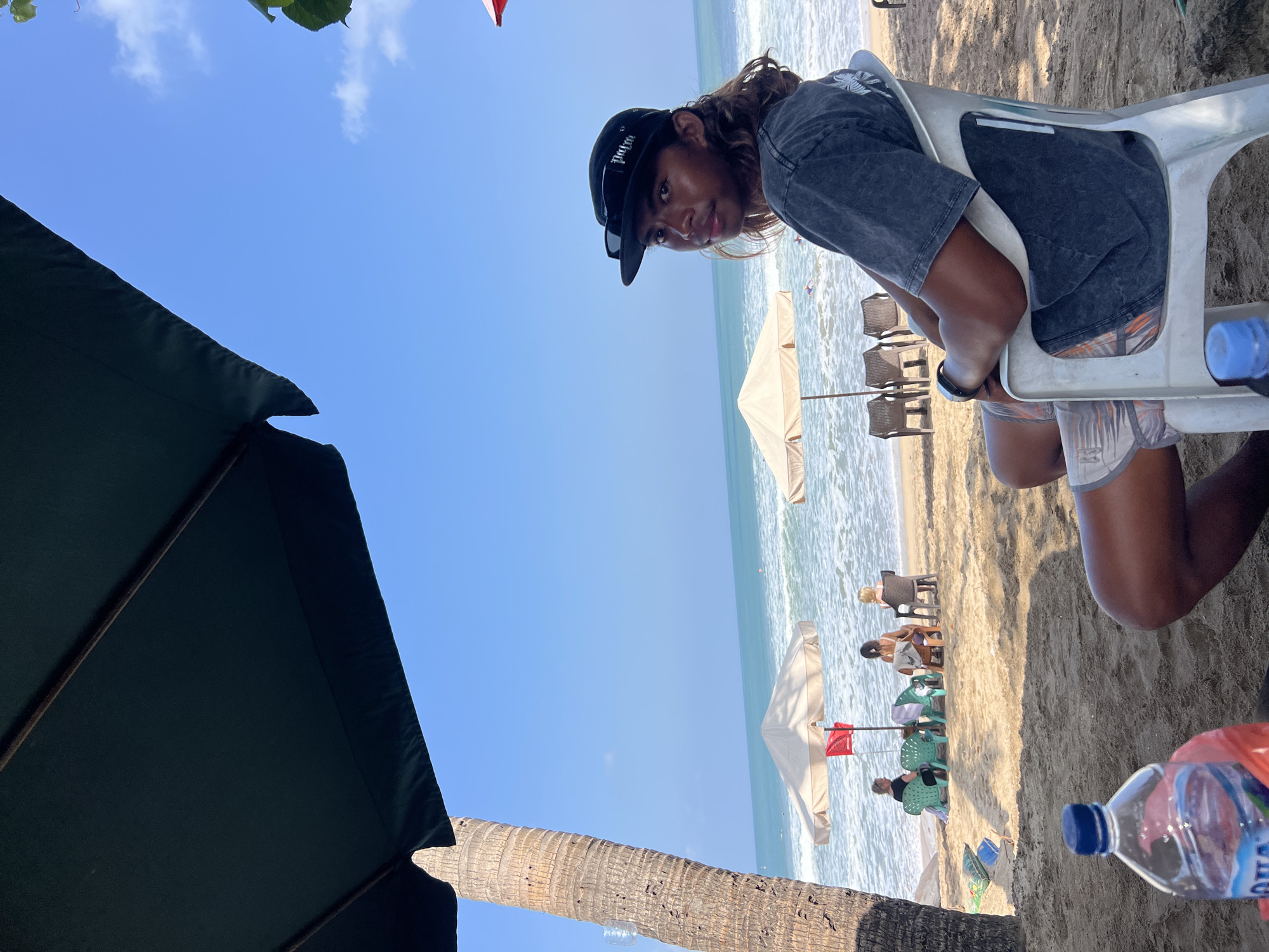 A young man with long, wavy brown hair, wearing a grey t-shirt and patterned shorts, sits in a white plastic chair on a sandy beach in Kuta, Indonesia. He is looking to his right, with a black baseball cap and sunglasses on his head. In the foreground, a bottle of water rests on the sand. In the mid-ground, several beach umbrellas provide shade over empty chairs. The ocean stretches out to the horizon under a clear blue sky, with small waves breaking on the shore. Palm trees are visible on the left side of the image.
