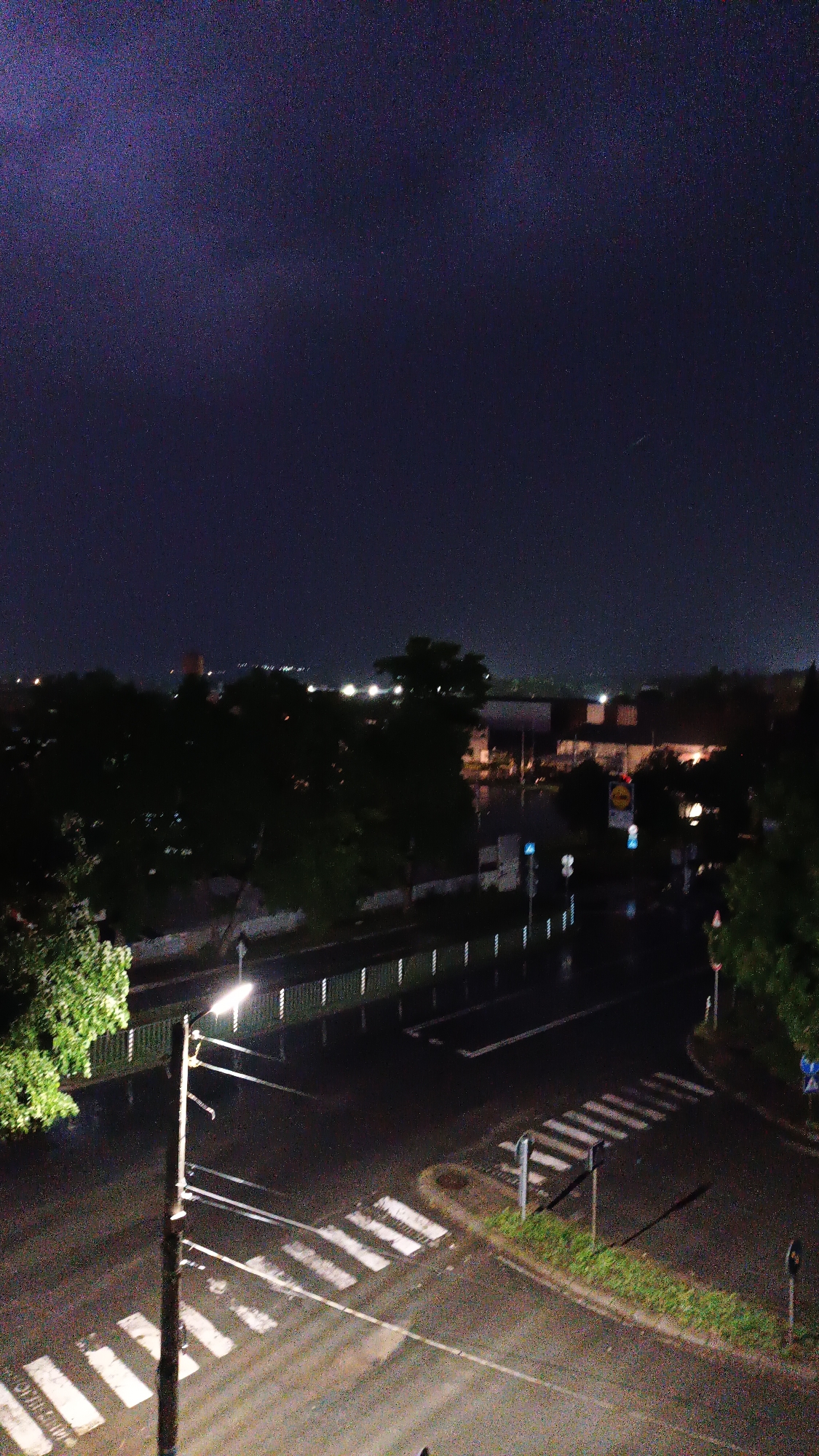 Here is a detailed description of the image:
The image depicts a night scene in an urban environment. The foreground consists of a wet asphalt street illuminated by a street light. A pedestrian crossing is visible, marked by white stripes. A lamppost is located nearby, with wires extending towards the street.
In the background, there are trees and buildings, with lights visible in the distance. A sign for Lidl is discernible. The sky is dark, with stars visible. The overall atmosphere suggests it is late at night after rain.