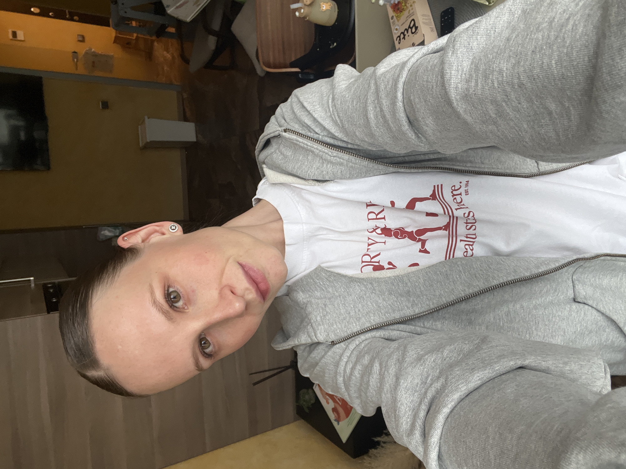 The image is a selfie of a young woman taken indoors, likely in a residence in Khimki, Russia. The woman, the primary subject, has dark hair neatly pulled back from her face, fair skin, and light brown or hazel eyes. She looks directly at the camera with a neutral to slightly content expression, her head tilted slightly.
She is wearing a casual outfit consisting of a light grey full-zip hooded sweatshirt layered over a white t-shirt. The white t-shirt features a red graphic design with stylized figures and fragmented text including "ORY & R", "calt s", "here.", and "EST. 1994.". On her left ear, she has two small silver earrings: a stud on the lobe and a small, circular earring higher on the cartilage.
The background reveals an indoor setting with warm-toned walls, possibly mustard yellow or light brown, and darker wooden furniture or panels. A dark rectangular object, possibly a television or a large framed picture, is visible on the left wall. A white rectangular object is mounted above it, and another white box-like item sits on a surface behind the woman. A box with the text "Bite" is partially visible on a counter or shelf in the background, alongside a bottle. The lighting suggests an indoor setting, likely with artificial light or diffuse natural light. The scene depicts a casual, everyday moment.