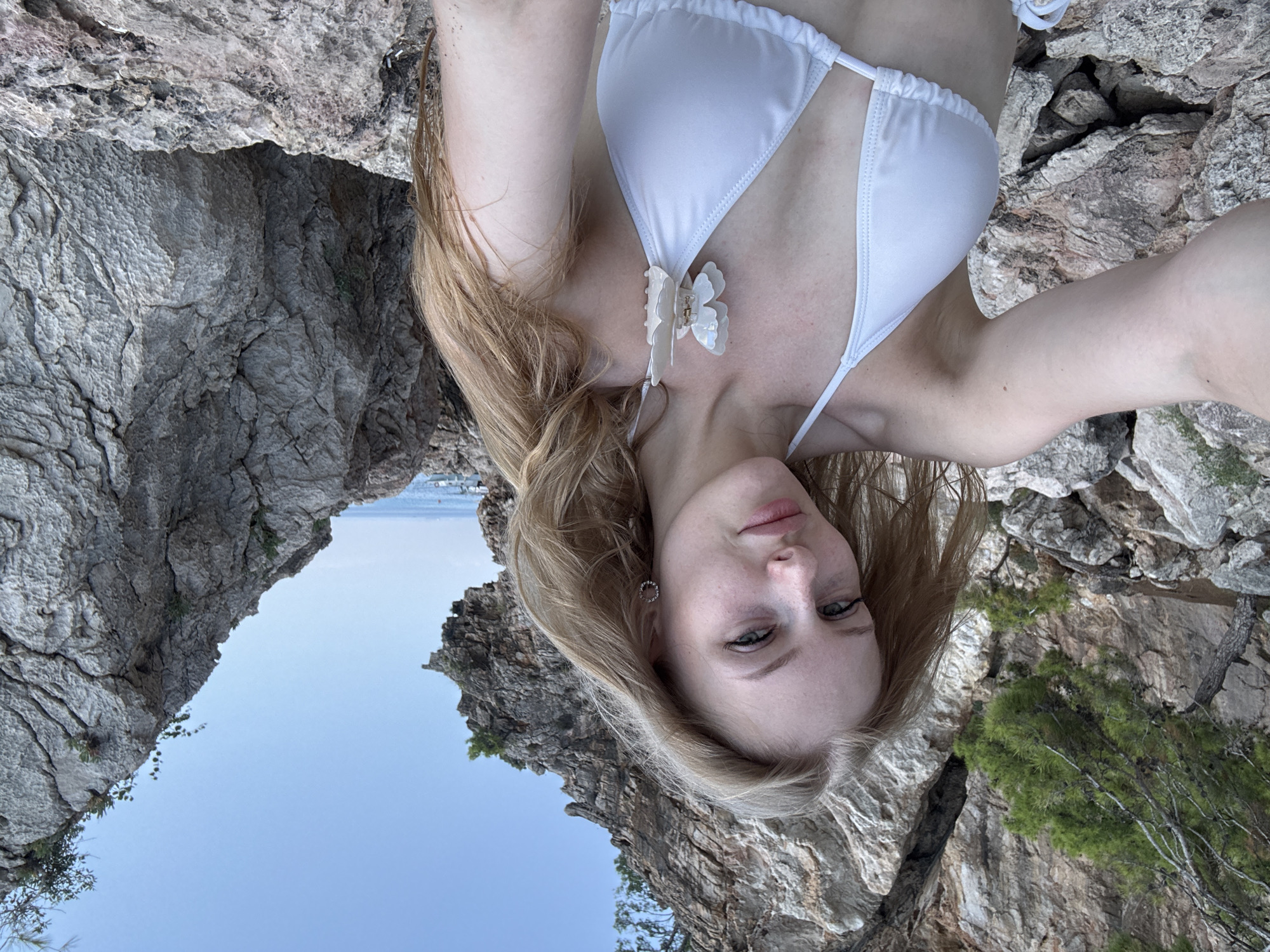 A young woman with long blonde hair is taking a selfie outdoors. She is wearing a white bikini top, adorned with a white butterfly clip on the left strap. She has light skin with freckles and is wearing subtle eye makeup. She is surrounded by a rocky, natural environment with large grey rocks and some greenery, likely on a coastline. In the background, the blue sea and sky are visible. The setting appears to be a natural coastal area, possibly a beach or cliffside, with a rugged landscape.