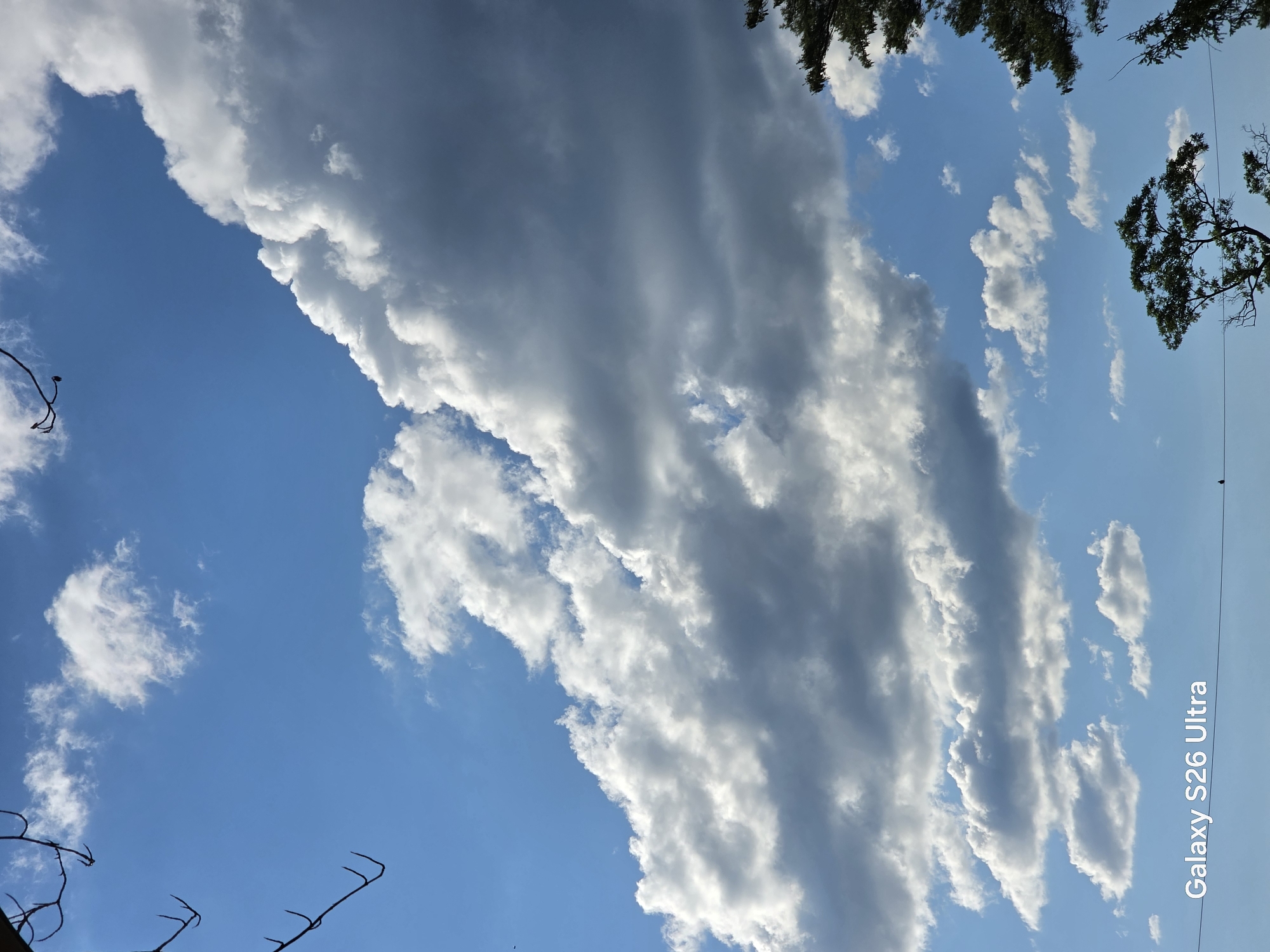 The image displays a bright blue sky filled with a mix of fluffy white and dark gray clouds. The clouds are dramatically arranged, with a large band of darker clouds dominating the upper right portion of the frame, casting shadows on the lighter clouds below. In the bottom right corner, the dark green foliage of a tree is visible, along with a portion of another tree in the upper right. The lower part of the image shows a thin black wire stretching horizontally across, with a small dark bird perched on it towards the right side. In the bottom left corner, the text "Galaxy S26 Ultra" is superimposed on the image. The scene is set outdoors, with the sky and clouds suggesting a dynamic weather pattern, possibly on the outskirts of West Hartford, Connecticut.