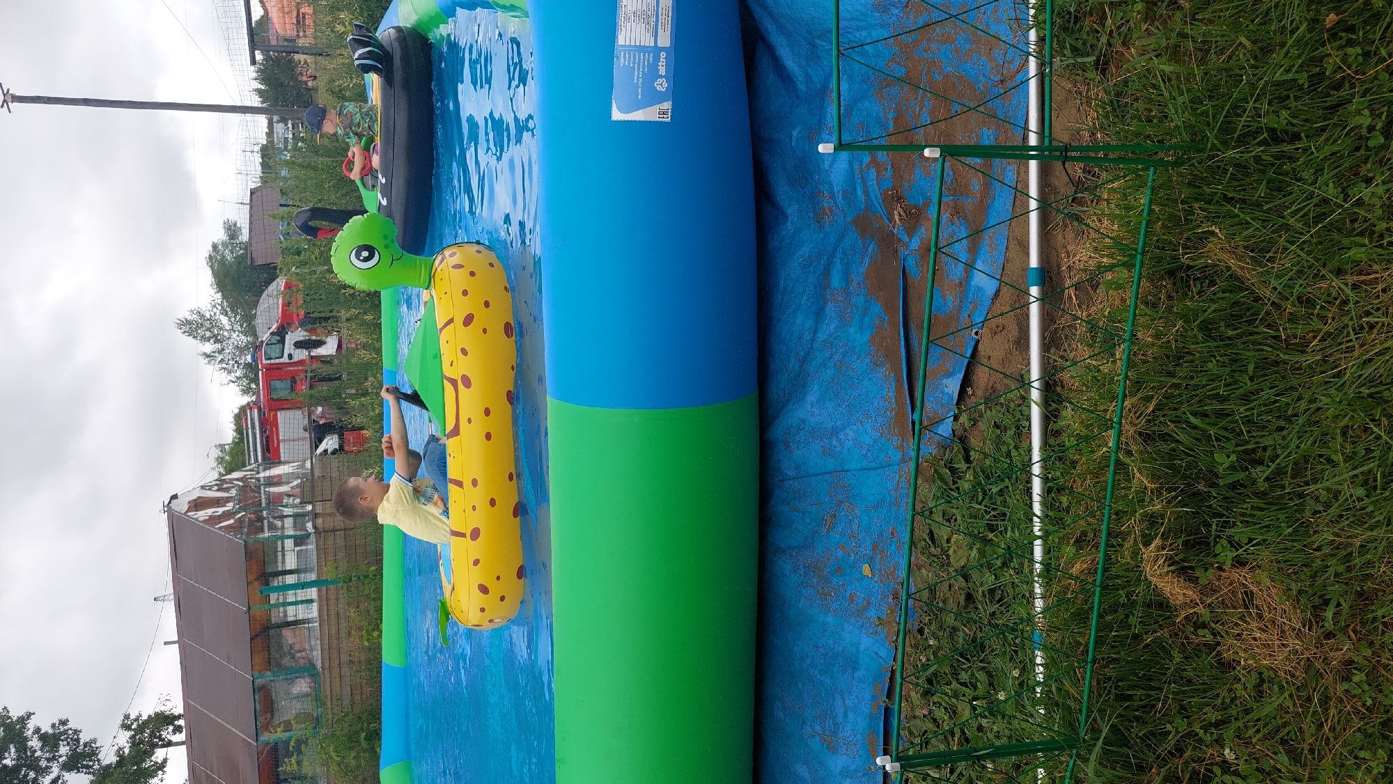 In Komsomolsk-on-Amur, Russia, a boy in a yellow shirt and blue jeans is seated in a yellow inflatable raft with red spots, holding a black steering wheel. He is in a large, blue and green inflatable swimming pool filled with clear blue water. To the right of the yellow raft, another inflatable raft, black with white accents and resembling a swan, carries a boy wearing a blue cap and a camouflage shirt. He is also holding a steering wheel. In the background, a red fire truck is visible, along with buildings and trees under a cloudy sky. The swimming pool is set up on a blue tarp, and a green metal fence with a diamond pattern encloses the area, with grass and dry vegetation in the foreground.