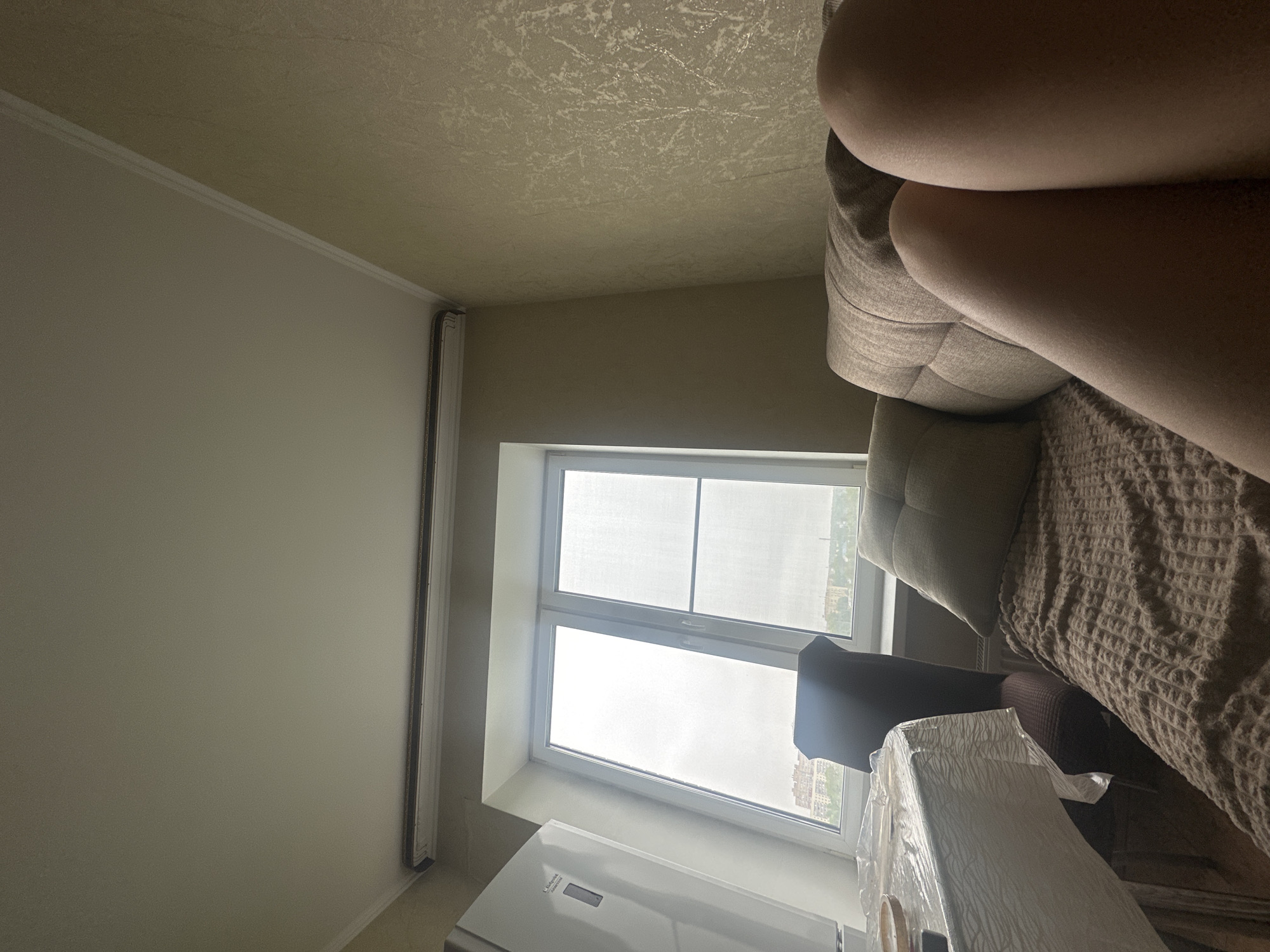 A domestic interior includes a window, a partially visible refrigerator, and a covered object, potentially furniture. A sofa, occupied by a person (identified by their upper thighs), is also present. The view through the window shows buildings, suggesting an urban environment. While the image's details are limited and lack specific landmarks, identifying the precise location in Naro-Fominsk, Russia, from the visual data alone is impossible.