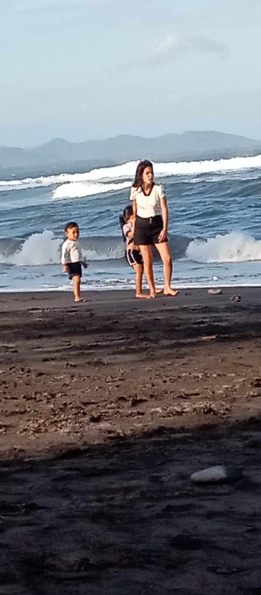 The image captures a beach scene in Blahbatuh, Indonesia, featuring three individuals. In the foreground, a young child stands on dark sand, looking towards the left. Slightly behind and to the right of the child is another child, partially obscured, wearing a black and white striped outfit. A woman, also on the sand, stands to the right of the children, looking towards the ocean. She is wearing a light-colored short-sleeved top and dark shorts. The ocean is visible in the background with moderate waves and white foam. Further in the distance, mountains are silhouetted against a hazy sky. The sand appears damp and textured, with some small pebbles and debris scattered on it.