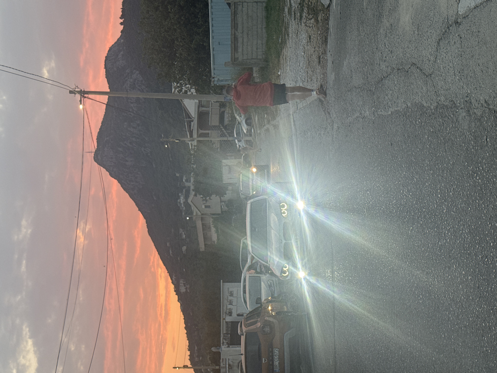 The image depicts a street scene in Sutomore, Montenegro, during either sunrise or sunset, with a vibrant orange and pink sky over a mountainous backdrop. In the foreground, a man wearing a red hoodie and black shorts is walking away from the camera on a paved road. The road appears wet, and there are several cars on it, some with their headlights on, causing lens flares. One car in the center is a white sedan with its headlights on. To the left, an orange SUV with license plate BR CC 672 is visible. Behind the cars, there are buildings and structures, some appearing residential. The environment suggests a somewhat unkempt or developing area, with a mix of construction materials and vegetation.